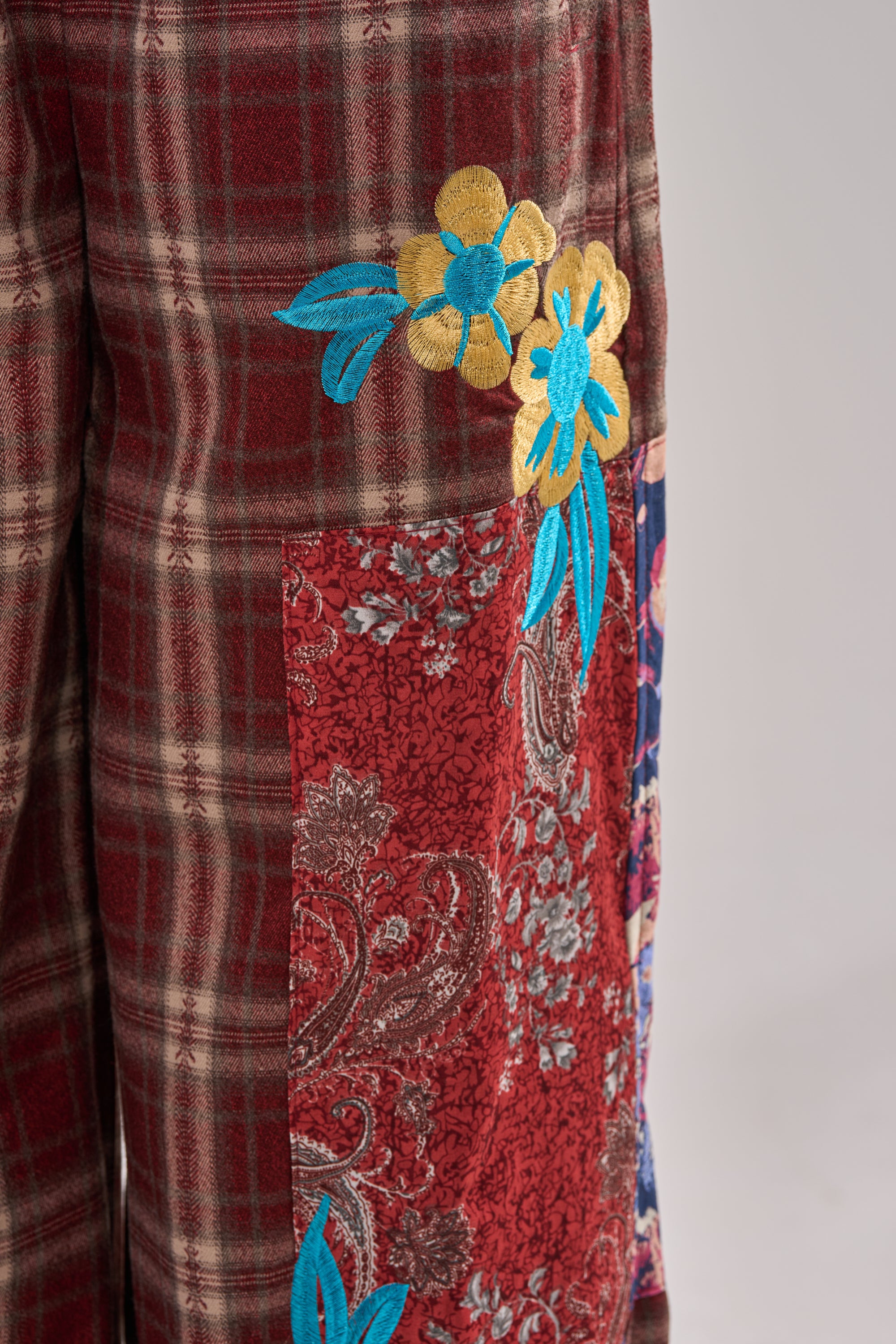 Close-up of the KRIYSTA FLORAL PATCH PLAID PANT, showcasing wide leg, high waist, brown and red plaid fabric, a red floral paisley patch, and embroidered yellow-blue flowers with blue leaves. 7