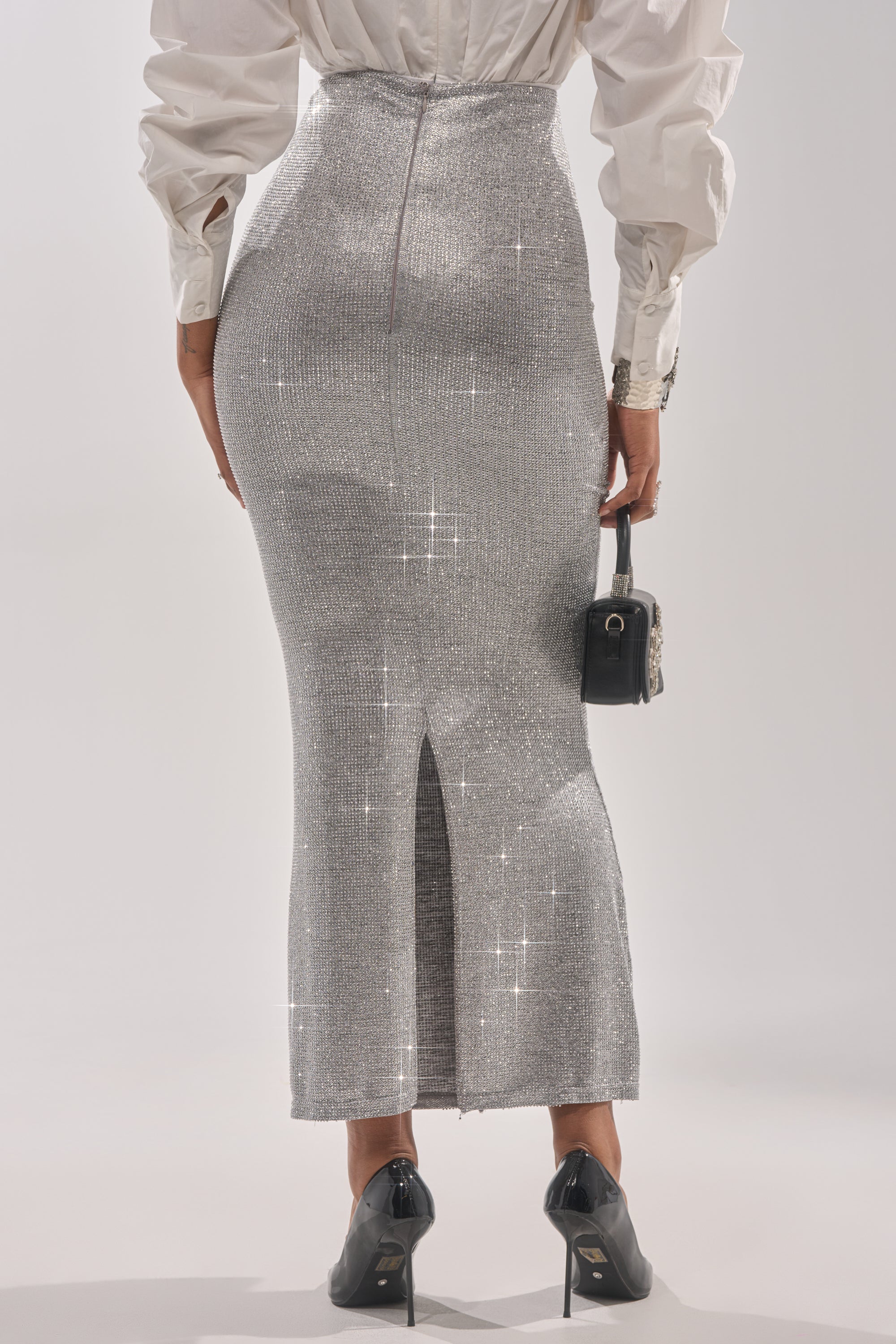 A woman faces away wearing the BRIGHT LIGHTS RHINESTONE MAXI SKIRT IN SILVER, paired with a white blouse, black heels, and a small black handbag—an elegant evening look set against a plain, light background. 3