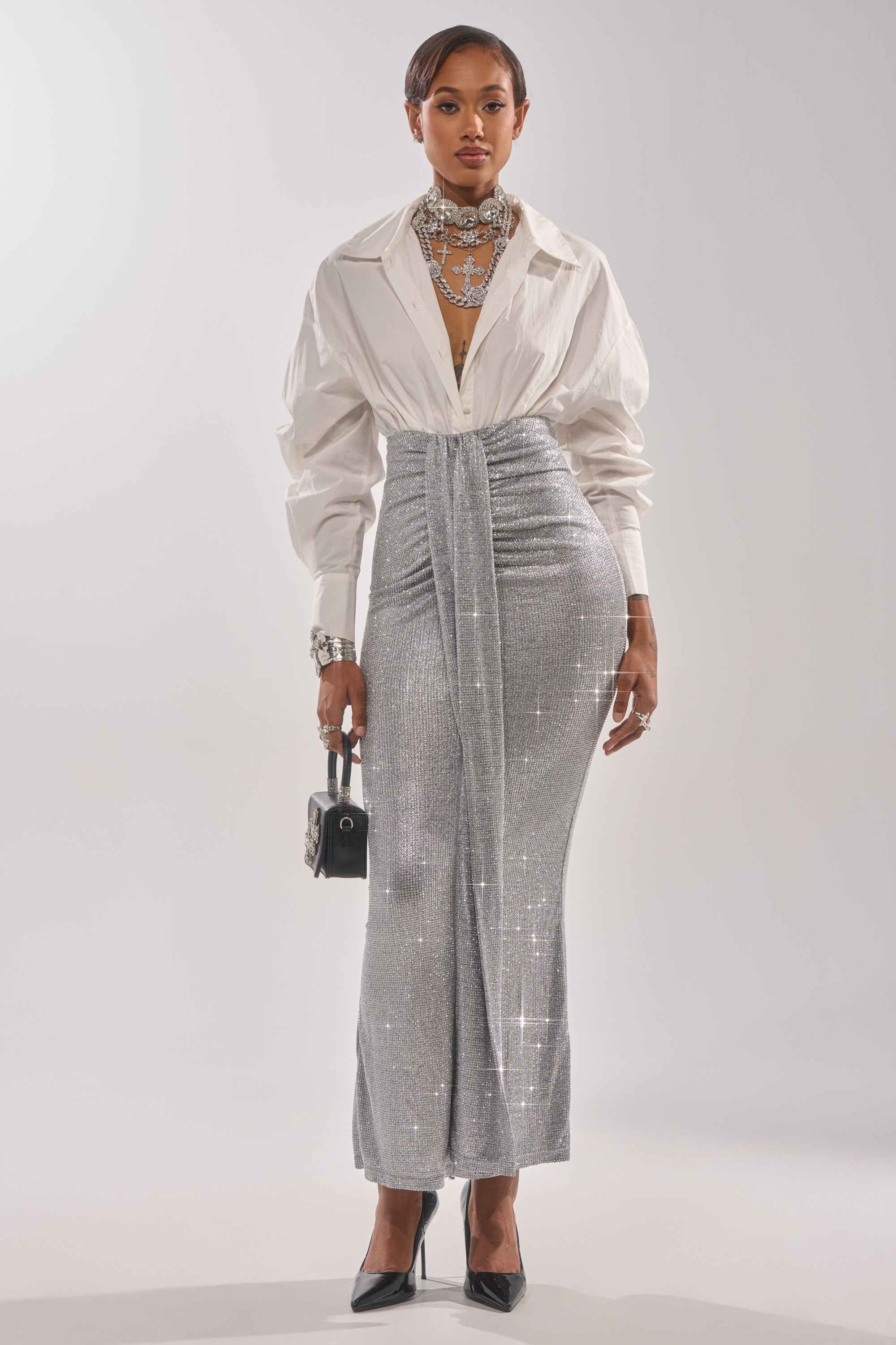 A woman faces forward, wearing the BRIGHT LIGHTS RHINESTONE MAXI SKIRT IN SILVER, a white deep-neck blouse, black heels, layered silver jewelry, and holding a small black handbag—an ideal chic look for evening events. 4