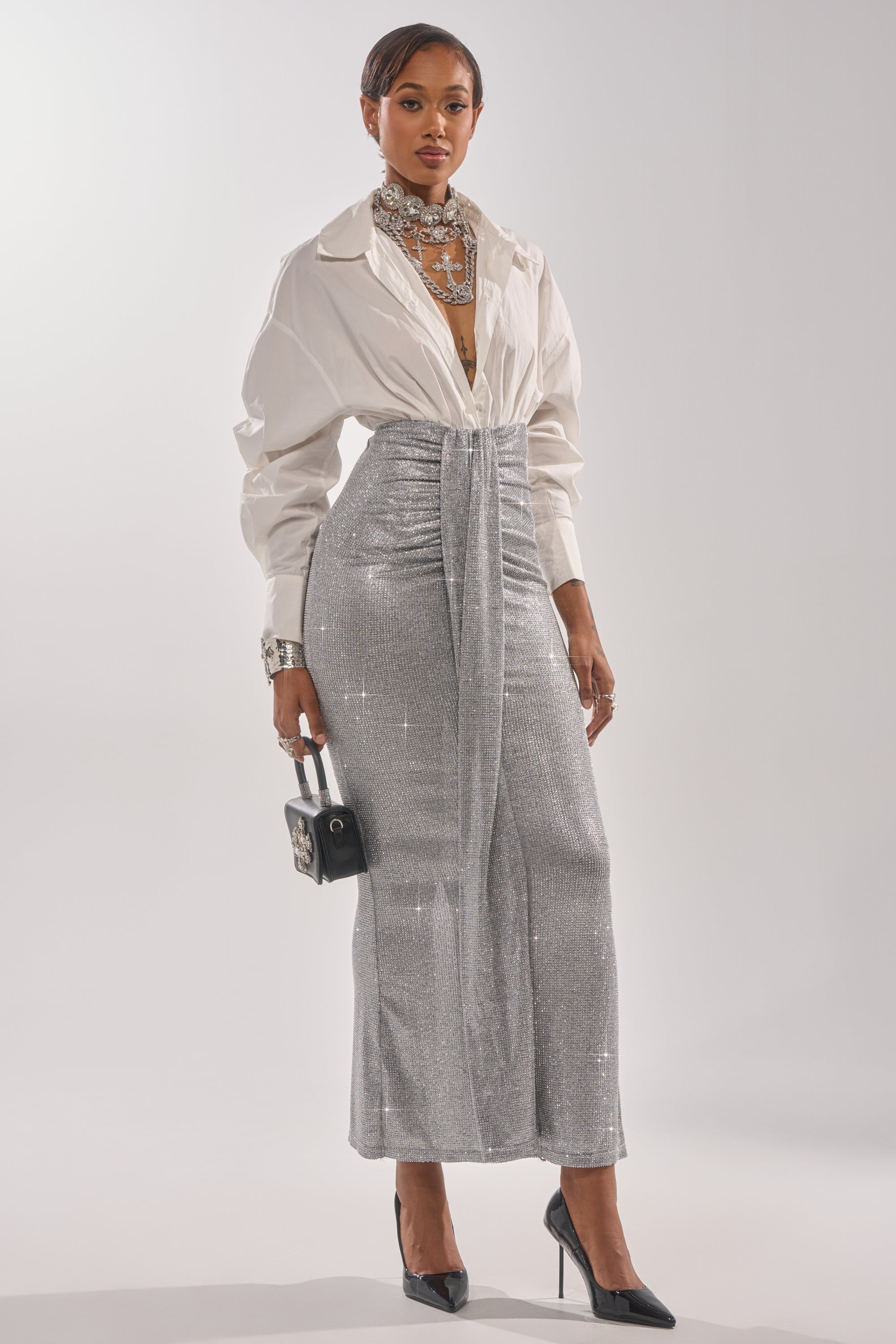 A woman models the BRIGHT LIGHTS RHINESTONE MAXI SKIRT IN SILVER with a white button-up, silver jewelry, black handbag, and pointed heels—a chic evening look highlighted by the skirt’s rhinestone shine and draped detail. 5