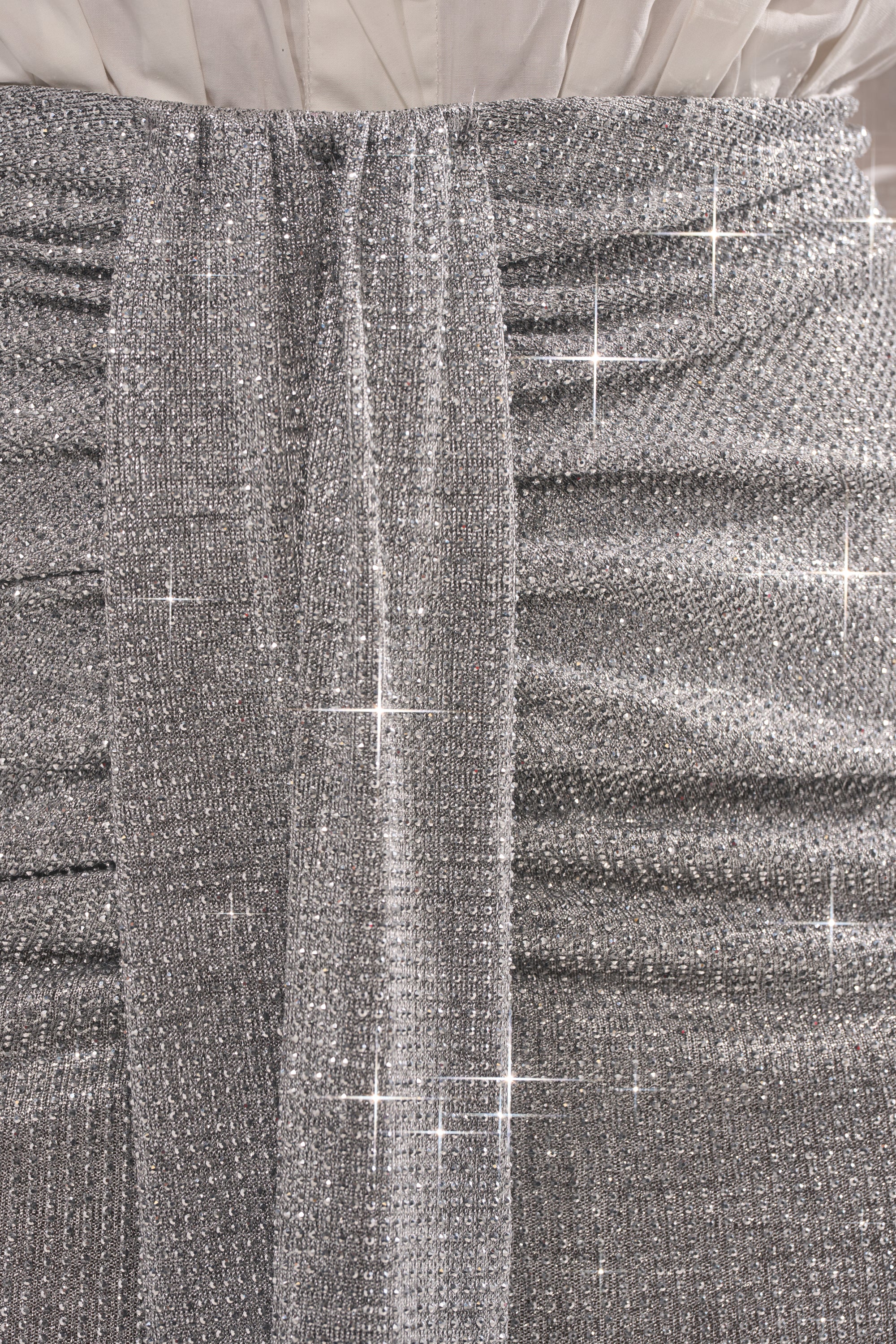 Close-up of the BRIGHT LIGHTS RHINESTONE MAXI SKIRT IN SILVER featuring a ruched design and shiny sash at the waist, styled with a white pleated top. The sparkling fabric glimmers, making it perfect for chic evening wear. 6