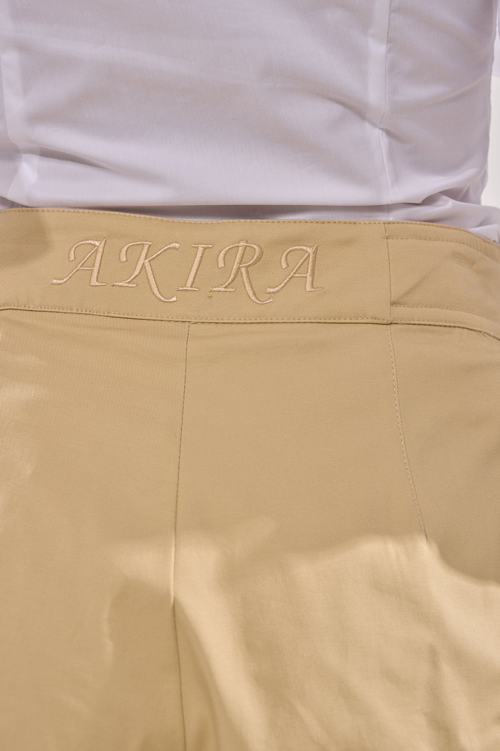 Close-up of the back of a person wearing beige SONIC PLEATED SHORTS with high waist, pleated details, and "AKIRA" embroidered in beige on the waistband. A white shirt is neatly tucked into the shorts. 8