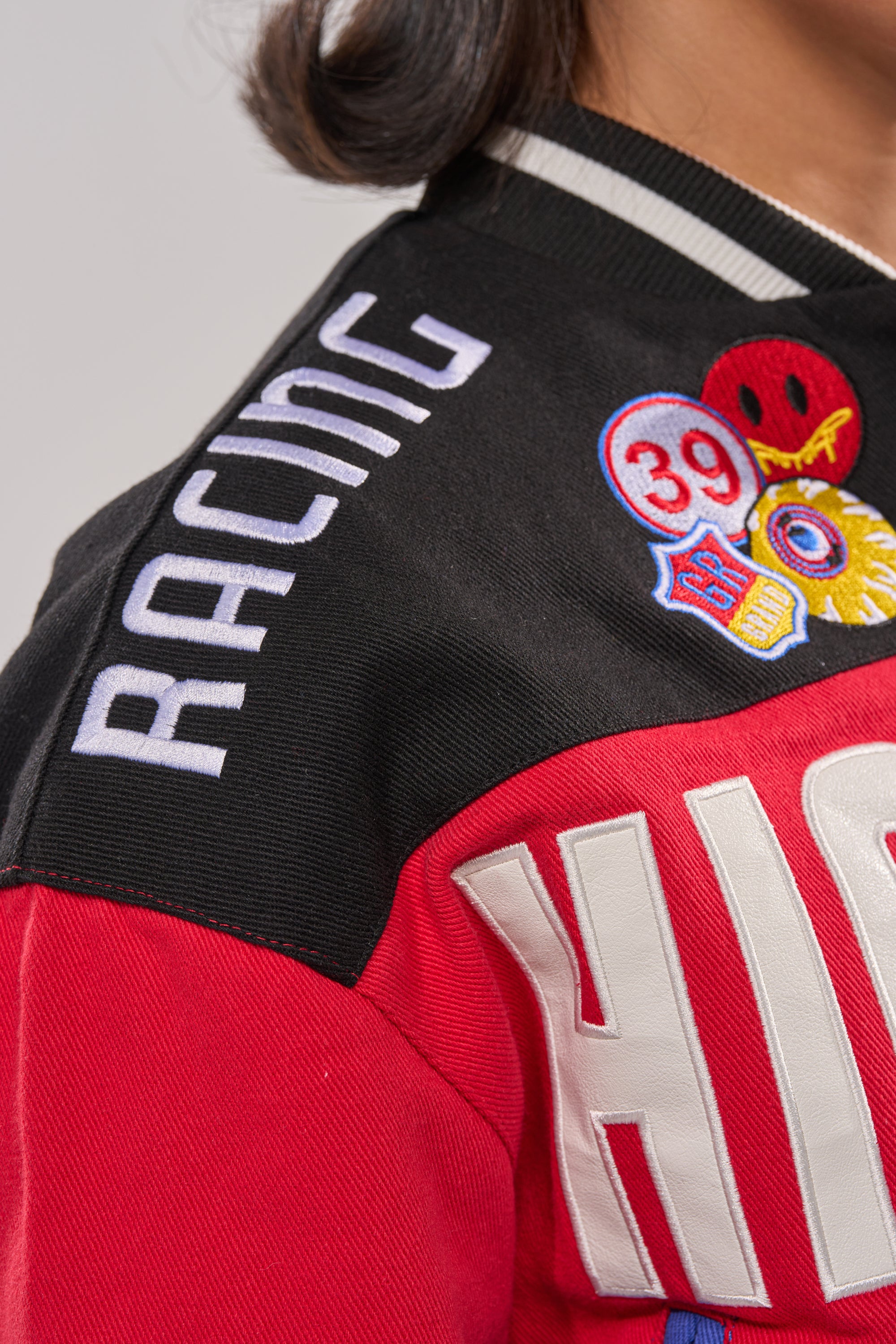 Close-up of a shoulder wearing the HIGH GEAR EMBROIDERED BOMBER, a colorblock varsity jacket with embroidered patches—a smiley face, "39," and "RACING" on the black sleeve.