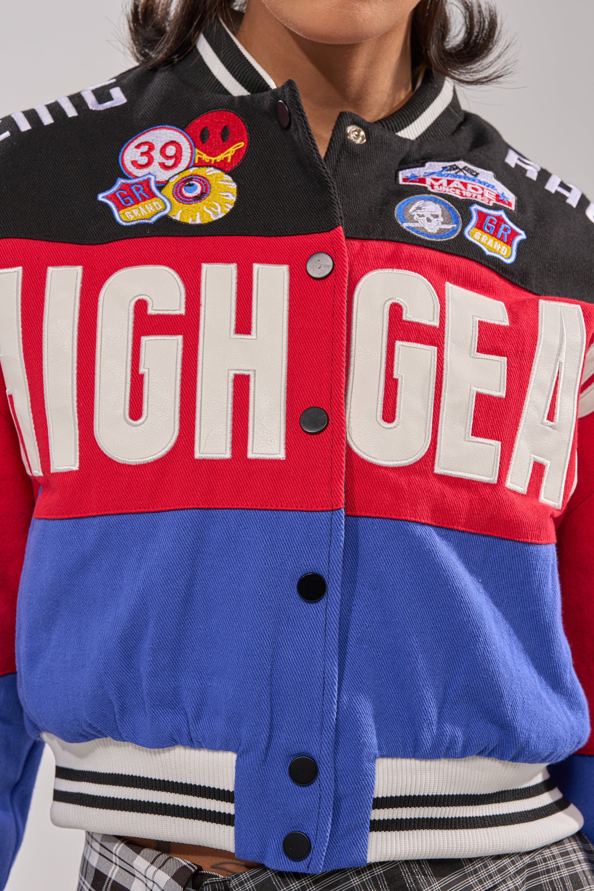 A cropped photo shows a person in the HIGH GEAR EMBROIDERED BOMBER, featuring bold "HIGH GEAR" text, decorative pins, and red, black, and blue colorblock patches, paired with black-and-white plaid pants.