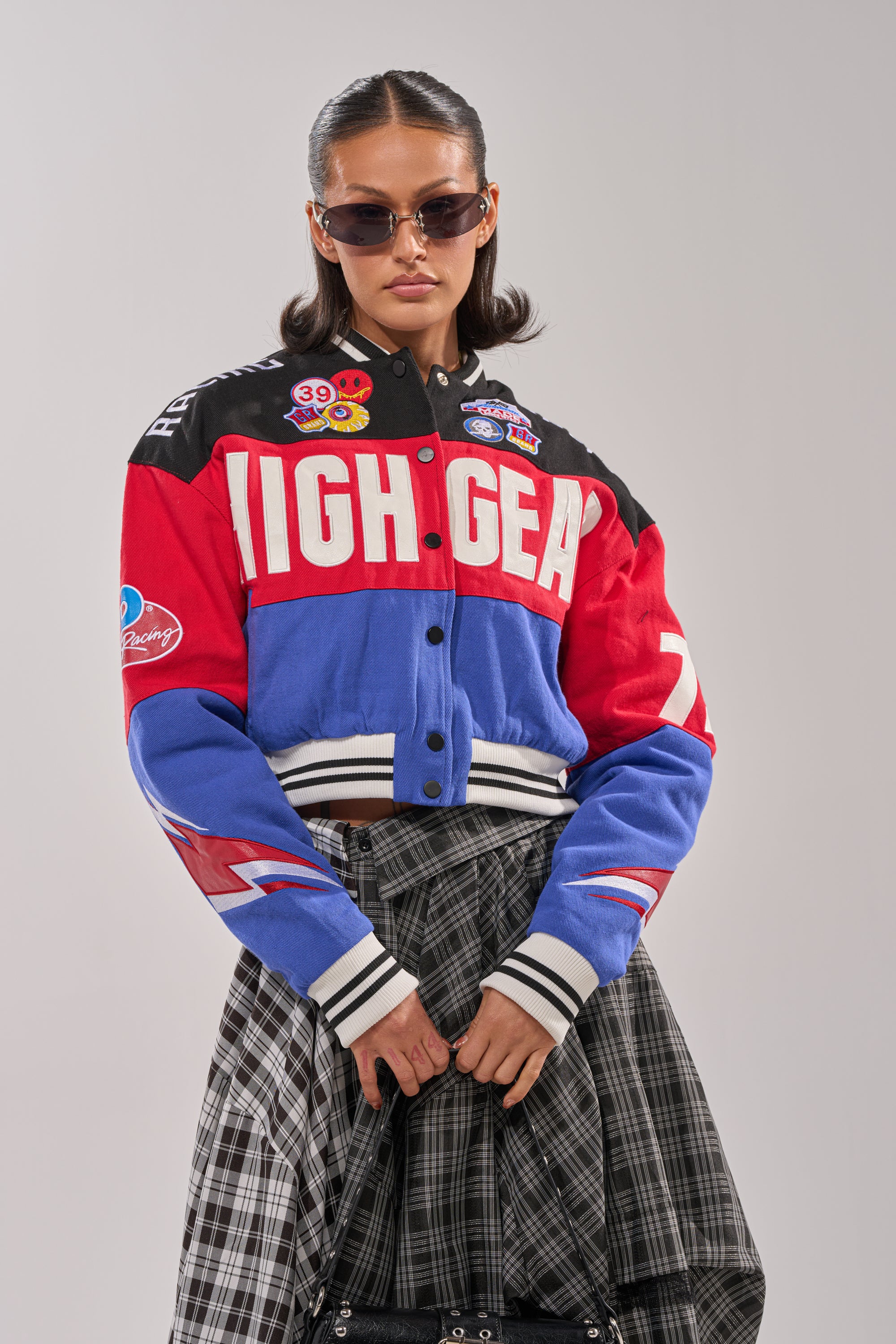 A woman wearing sunglasses and a colorful "HIGH GEAR EMBROIDERED BOMBER" with patches, paired with a black and white plaid skirt, stands against a light gray background.