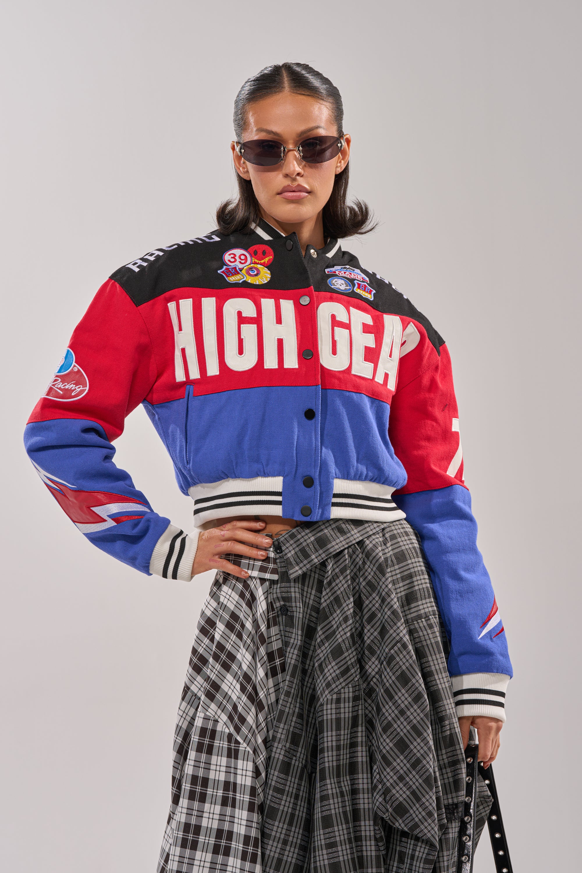 A woman in sunglasses and the HIGH GEAR EMBROIDERED BOMBER, a red and blue racing-inspired jacket with patches, poses confidently with one hand on her hip against a plain background.