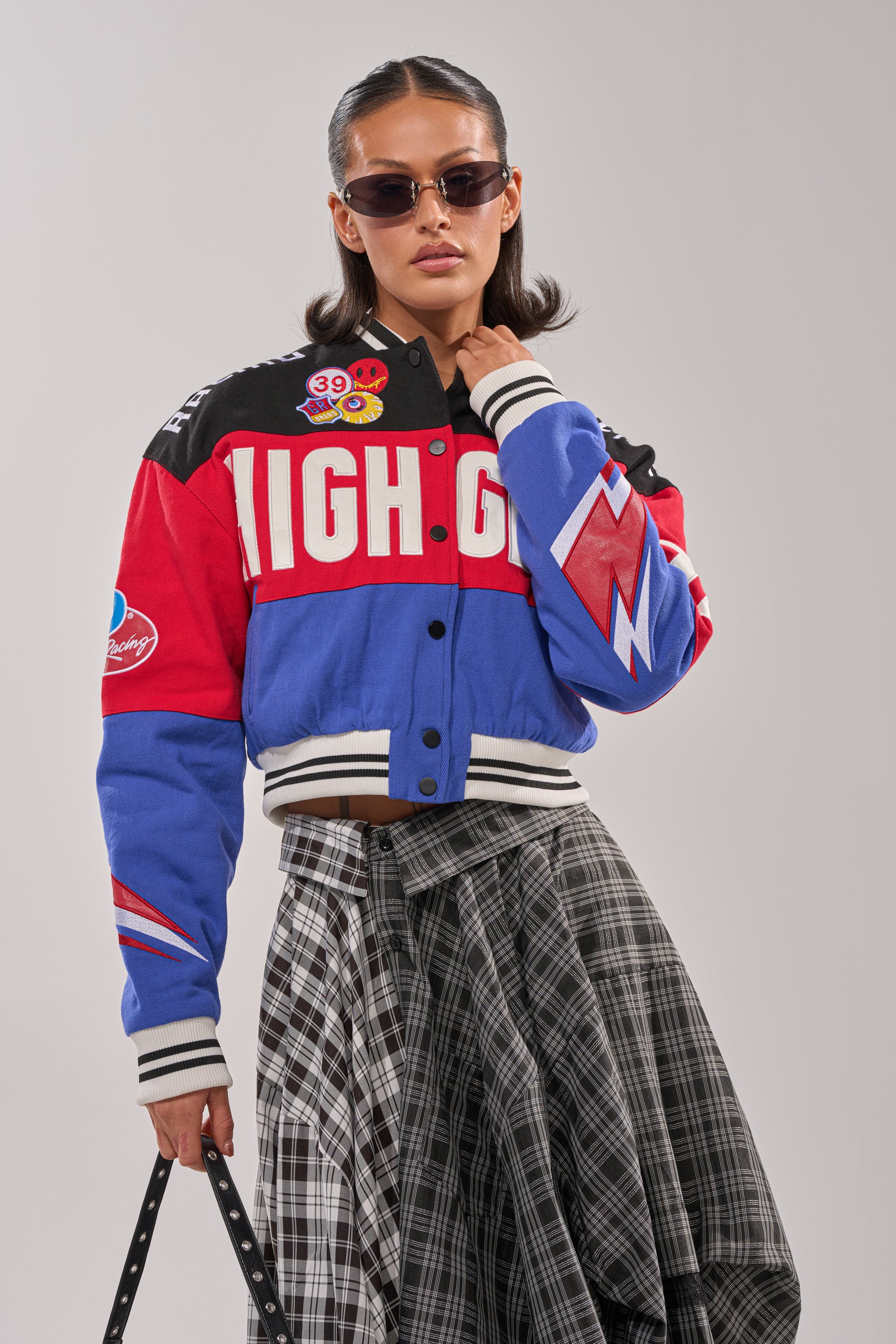 A person stands confidently against a plain background wearing sunglasses, a black-and-white plaid skirt, and the HIGH GEAR EMBROIDERED BOMBER— a colorful, racing-inspired varsity jacket.