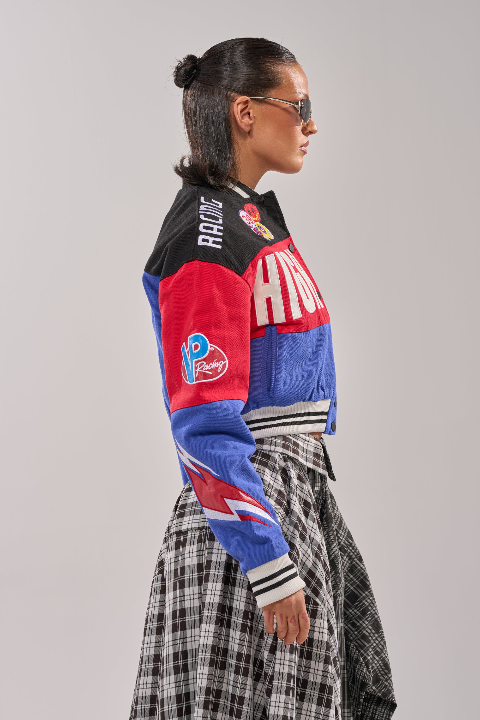 A woman with slicked-back hair and sunglasses models the HIGH GEAR EMBROIDERED BOMBER, featuring racing-inspired embroidery, paired with a black-and-white plaid skirt, shown in profile against a plain light background.