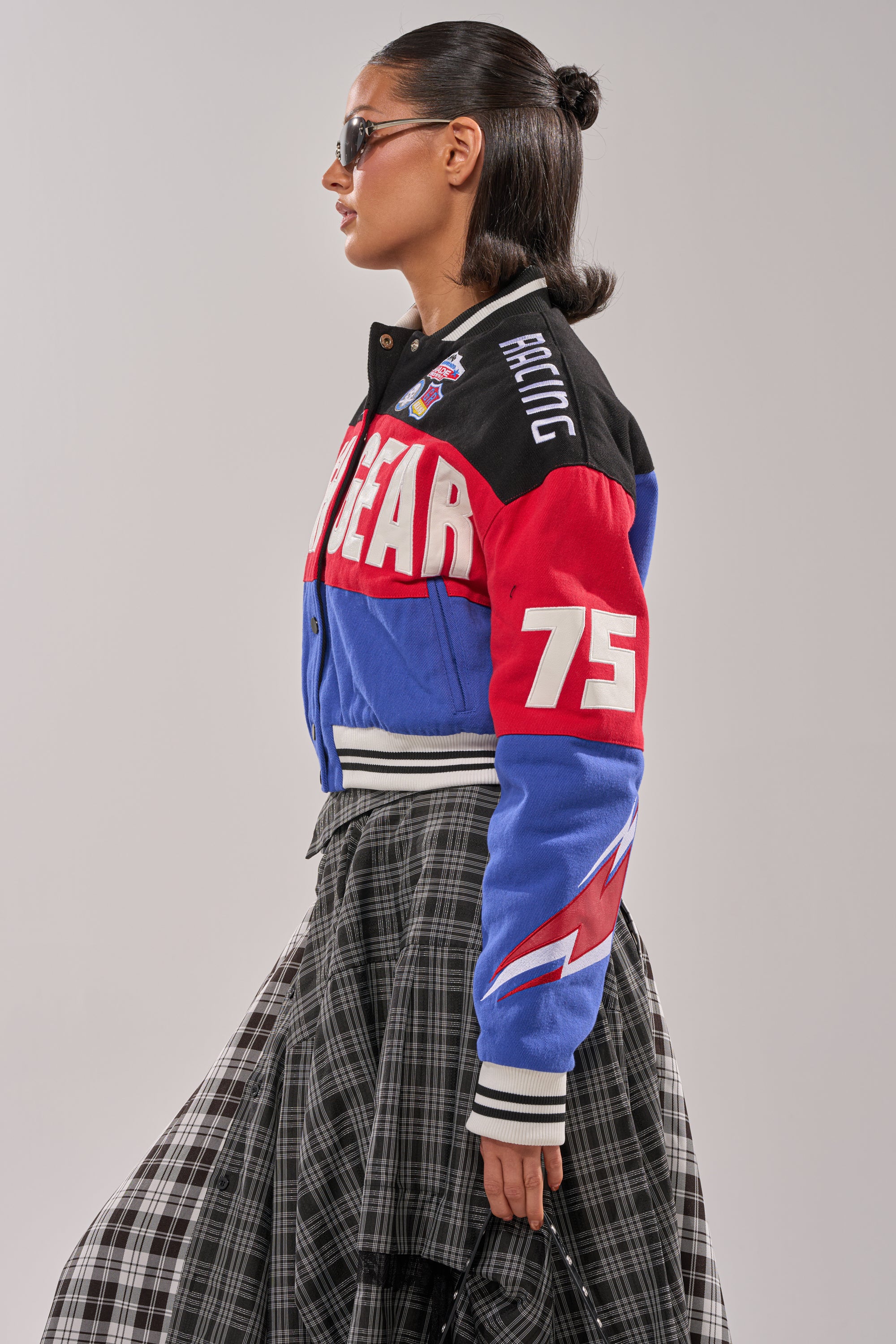 A woman in sunglasses wears the HIGH GEAR EMBROIDERED BOMBER, featuring colorblock panels, racing-inspired embroidery with "GEAR" and "RACING," paired with a pleated black-and-white plaid skirt. She stands sideways against a plain background.