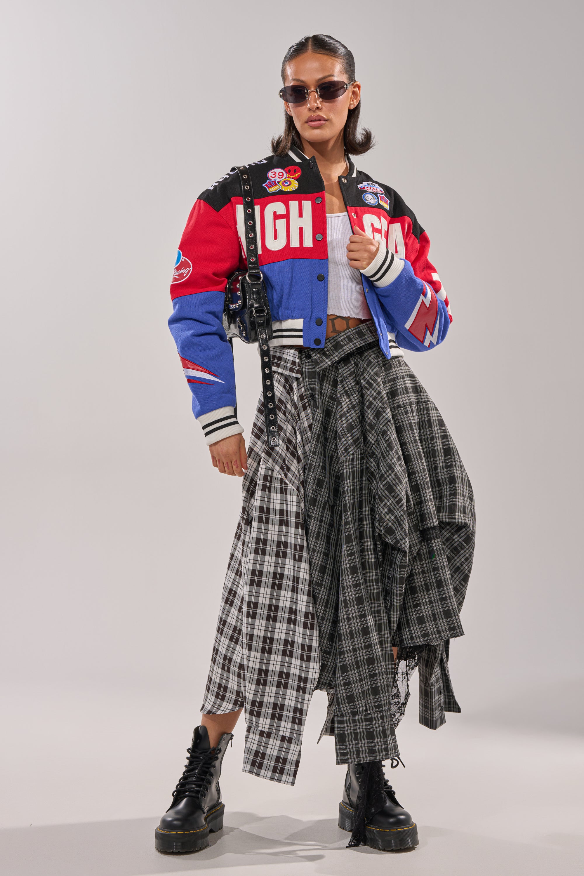 A woman stands confidently against a plain background, wearing sunglasses and the HIGH GEAR EMBROIDERED BOMBER with racing-inspired embroidery, paired with a gray crop top, black and white plaid skirt, and black platform boots.