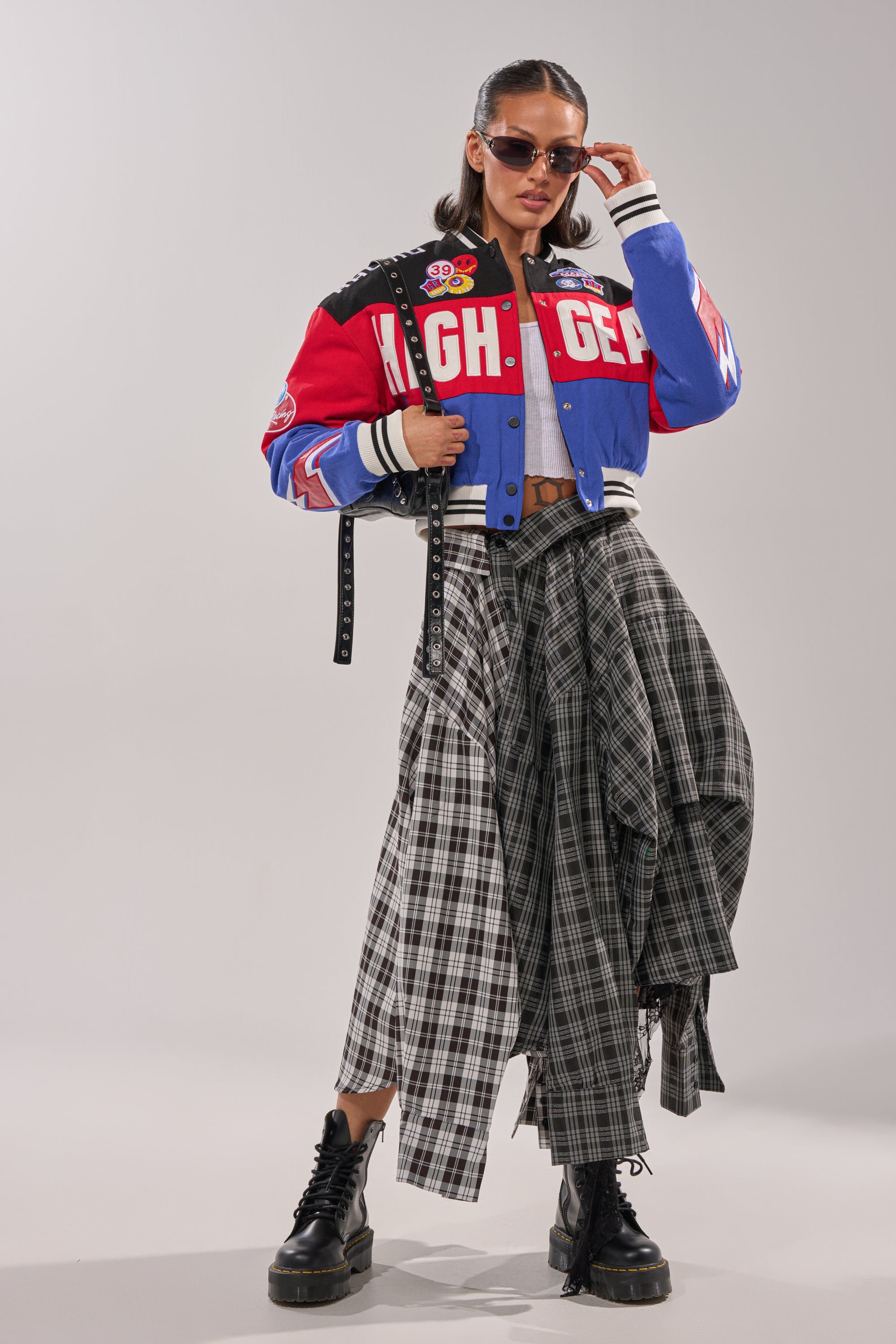 A woman poses confidently against a plain light background wearing sunglasses, chunky black boots, a long plaid skirt, black belt, and the HIGH GEAR EMBROIDERED BOMBER with colorblock style and racing-inspired details.