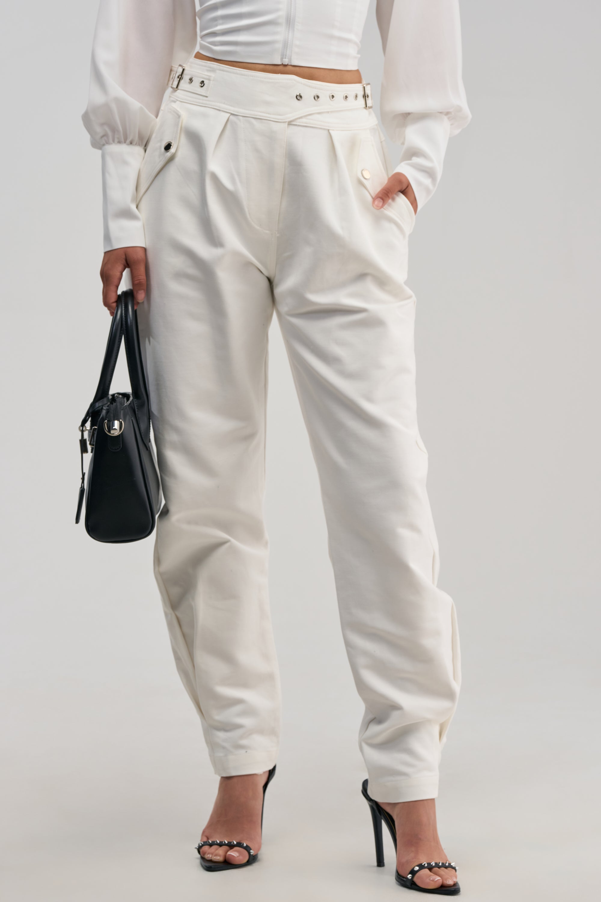 A person wearing the white HEY YOU TROUSER, paired with a white long-sleeve top and black high-heeled sandals, holding a black handbag. The photo captures the outfit from the shoulders down against a plain background.