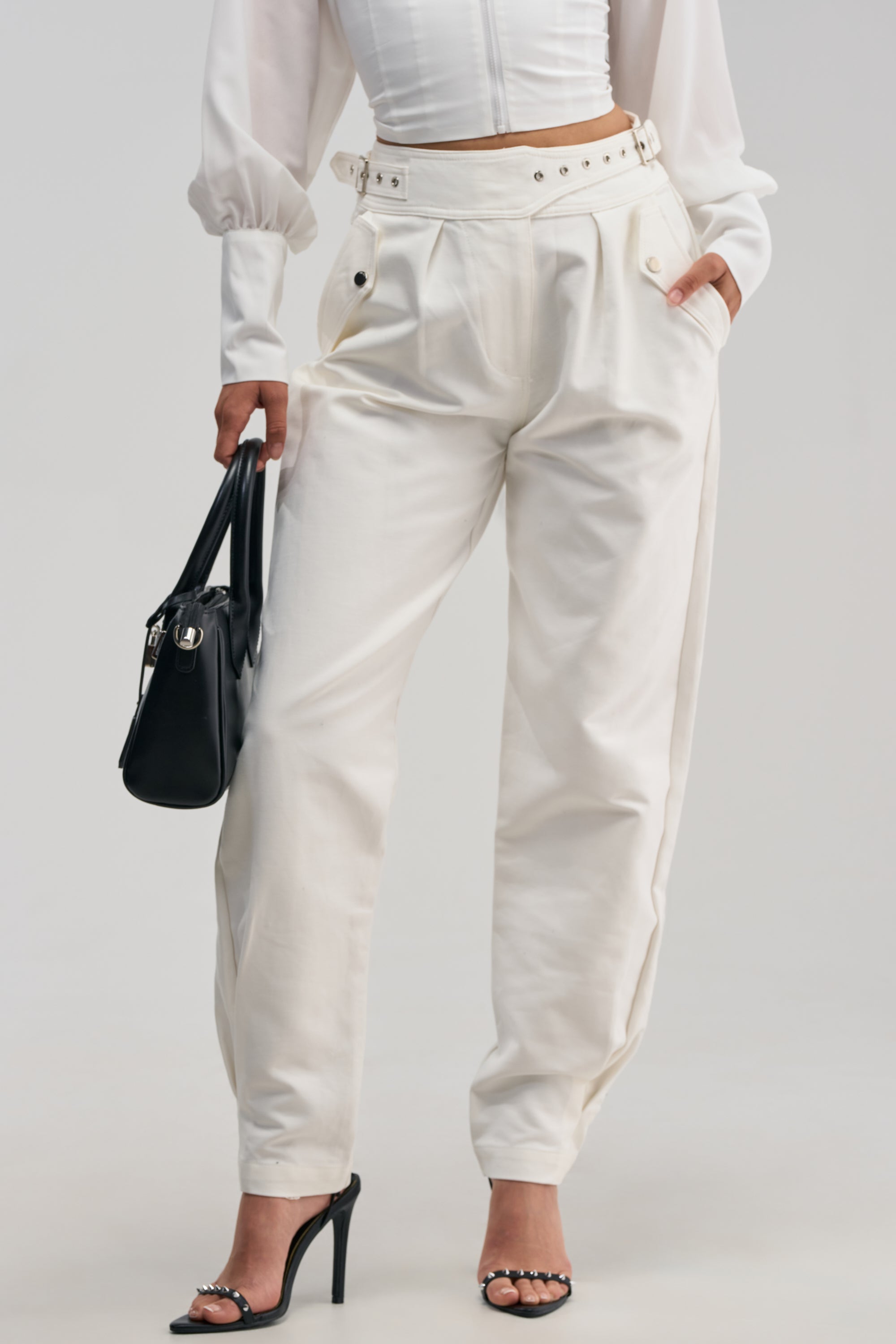 A woman is wearing the HEY YOU TROUSER, a pair of high-rise cotton trousers, styled with a white long-sleeve top, black high-heeled sandals, and carrying a black handbag against a plain light background.