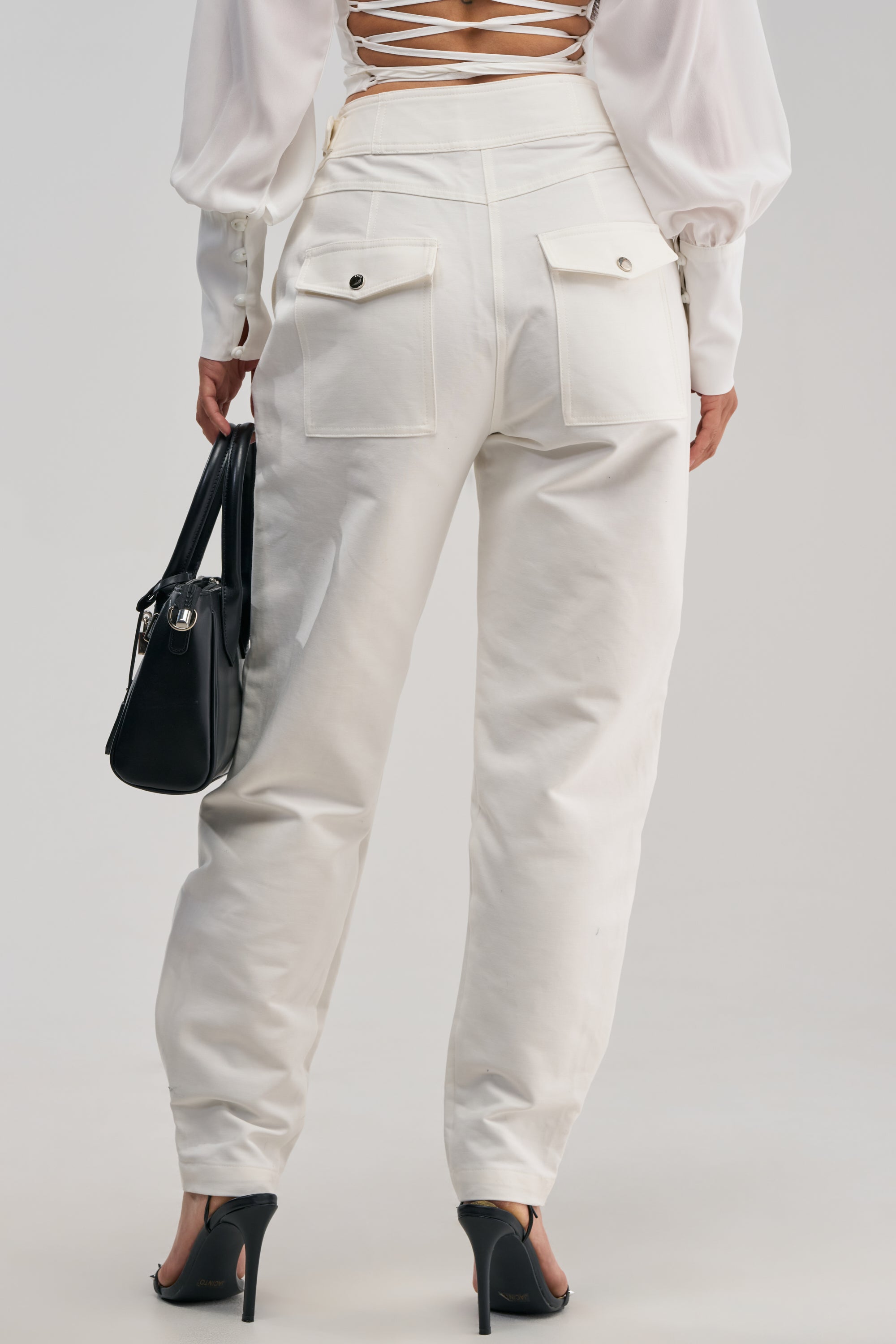 A person is shown from behind wearing the HEY YOU TROUSER, which are high-rise white pants with back pockets, paired with a white long-sleeve top featuring a lace-up back, black heels, and holding a black handbag.