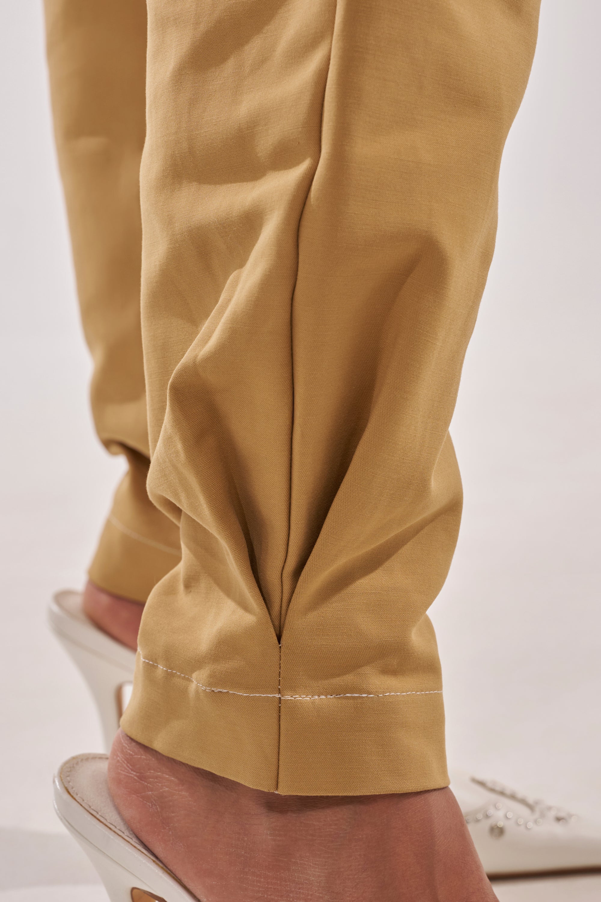 Close-up of a person's lower legs wearing the HEY YOU TROUSER, paired with open-toe white heeled sandals. The photo showcases the trouser's fabric and detailed stitching near the ankles. 9