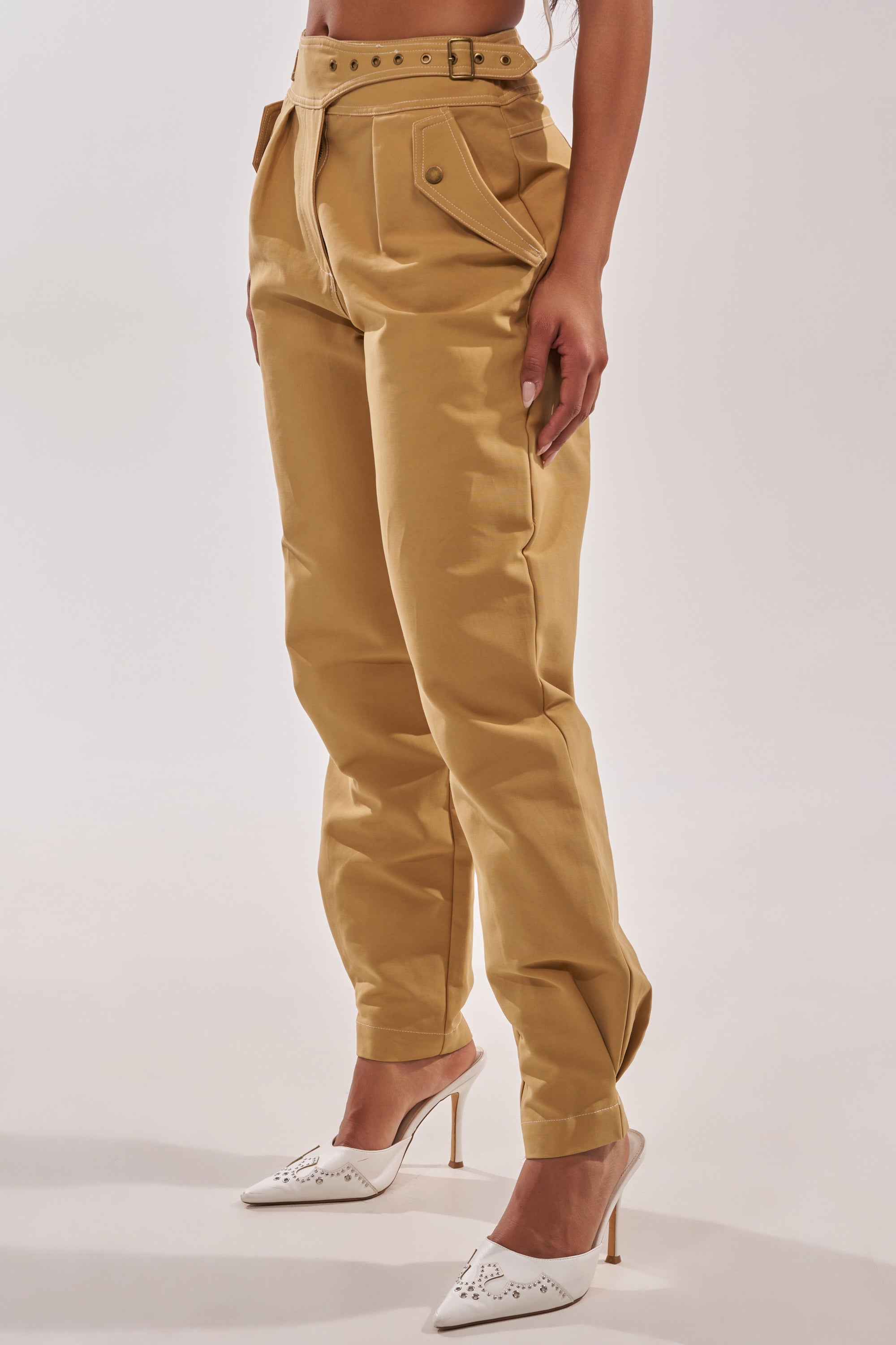 A person wears the HEY YOU TROUSER—high-waisted, tan utility-style pants with a belted waist—paired with white pointed-toe heeled shoes, against a plain light background. 2