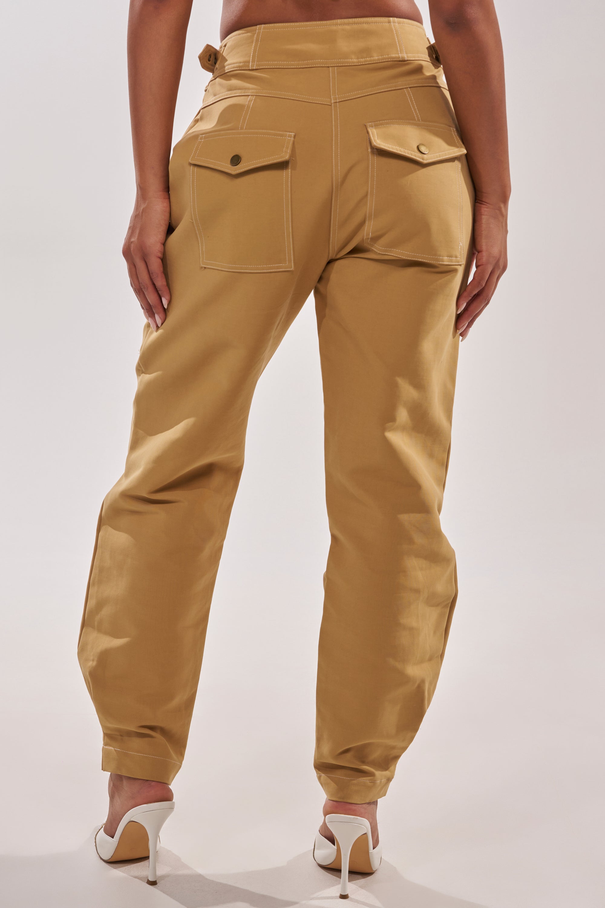 A person is seen from behind wearing the HEY YOU TROUSER in tan—a high-rise cotton style with large back pockets—paired with white high-heeled shoes, set against a plain light backdrop. 4