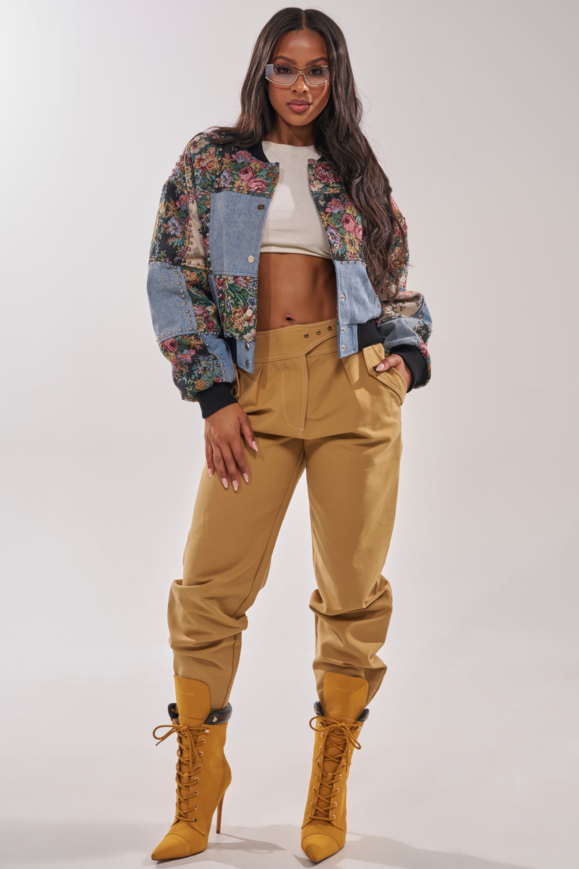 A woman with long wavy hair and glasses stands confidently with one hand in her pocket, wearing a patchwork denim and floral jacket, white crop top, HEY YOU TROUSER high-rise pants, and yellow heeled lace-up boots. 7