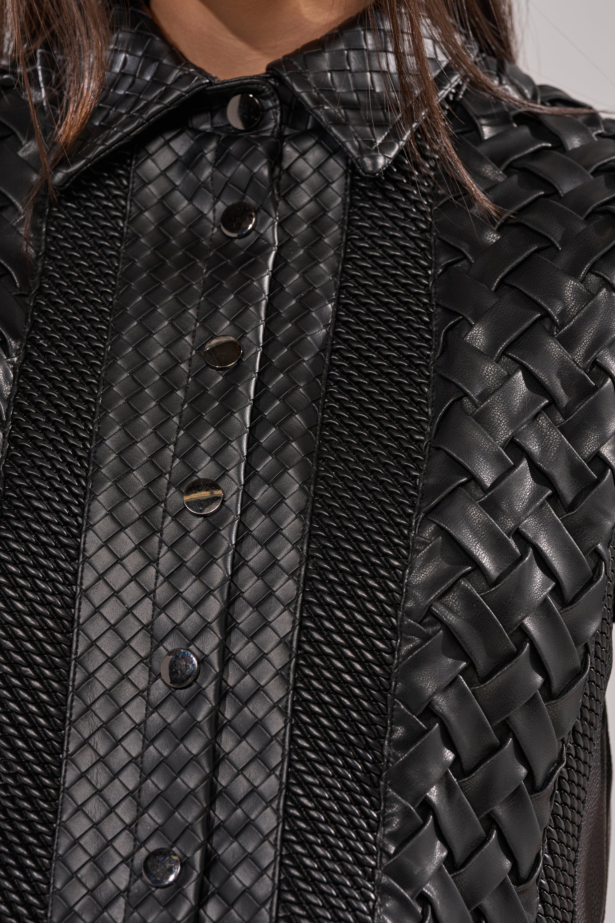 Close-up of the BIKER BRAIDED LEATHER CROPPED BUTTON DOWN, showcasing its black textured fabric with braided panels, smooth leather detailing, center buttons, and a visible collared neckline. 10
