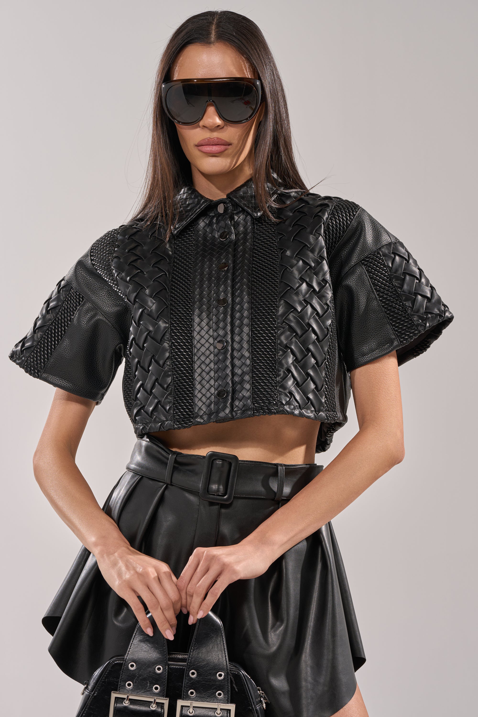 A woman wears the BIKER BRAIDED LEATHER CROPPED BUTTON DOWN with oversized black sunglasses, a belted black leather skirt, and holds a black purse against a plain background. 1