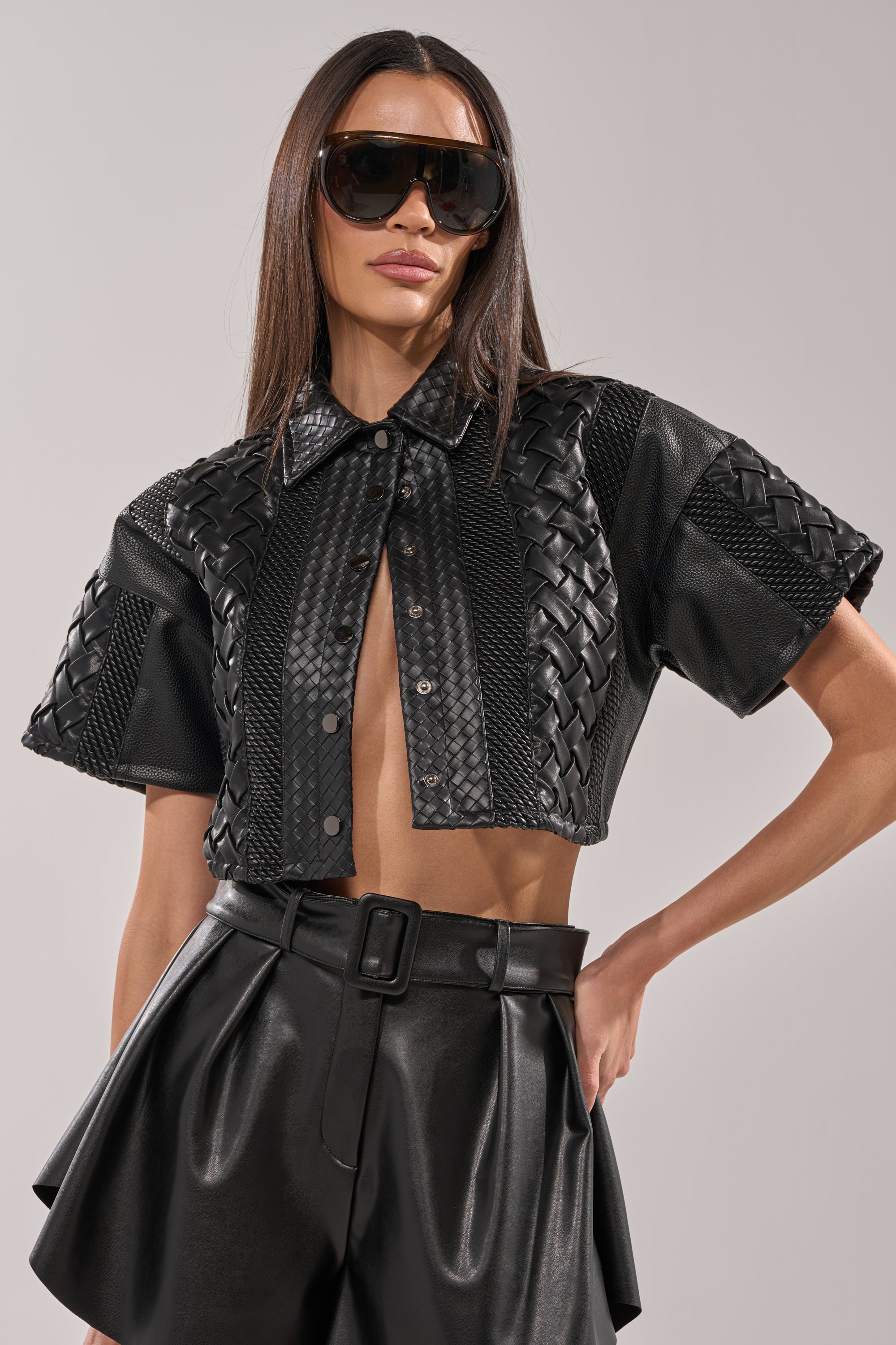 A woman poses confidently in the BIKER BRAIDED LEATHER CROPPED BUTTON DOWN—short-sleeved, collared, and paired with high-waisted black leather shorts and large sunglasses against a plain background. 3