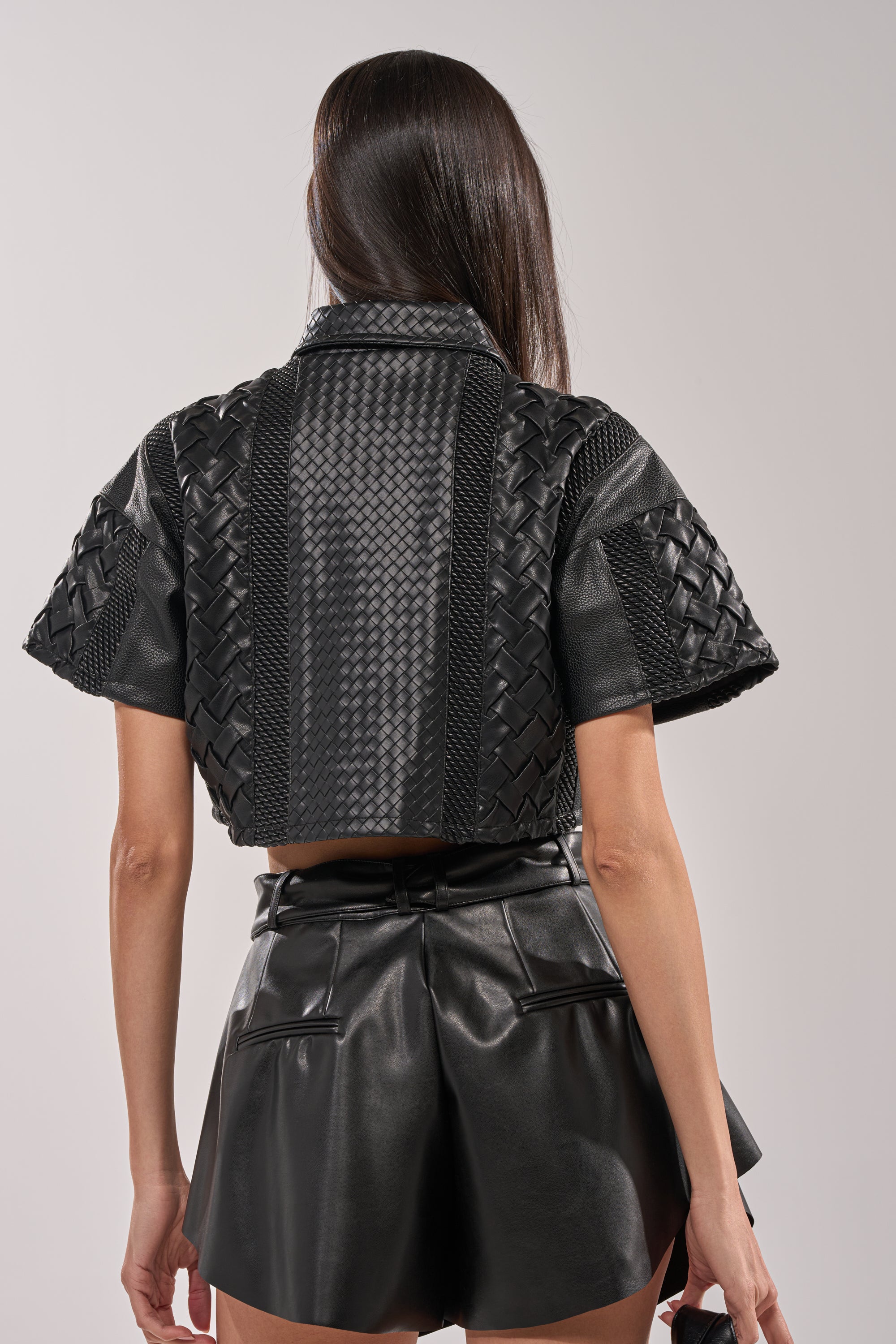 A person with long dark hair, seen from behind, wears an edgy women's look: the BIKER BRAIDED LEATHER CROPPED BUTTON DOWN paired with a high-waisted pleated faux leather mini skirt. 6
