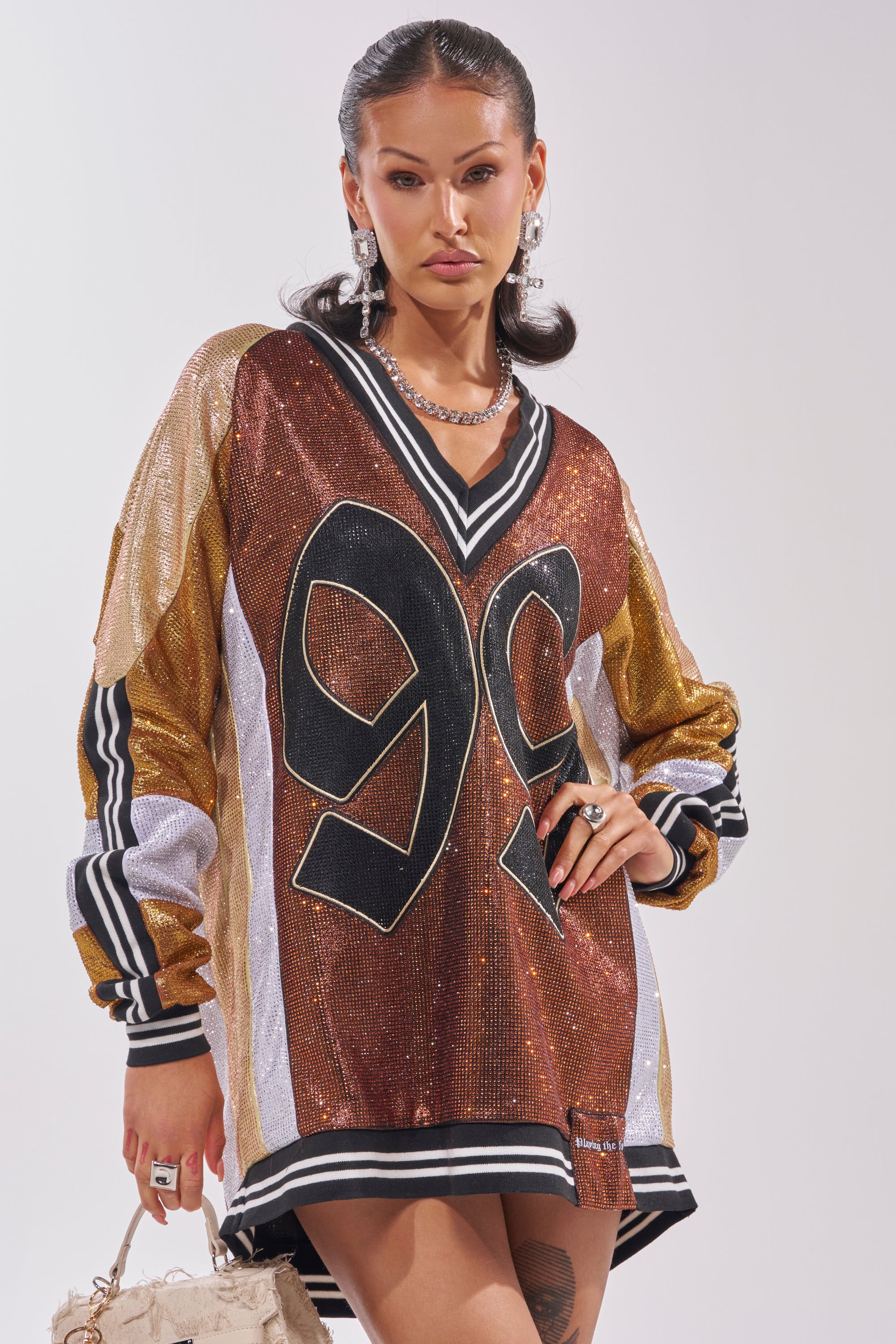 A woman poses confidently in the RYAN COLORBLOCK RHINESTONE JERSEY DRESS IN COCOA, an oversized sports-inspired dress with gold, bronze, and silver panels. She accents her look with chunky silver jewelry and a textured beige bag. 3