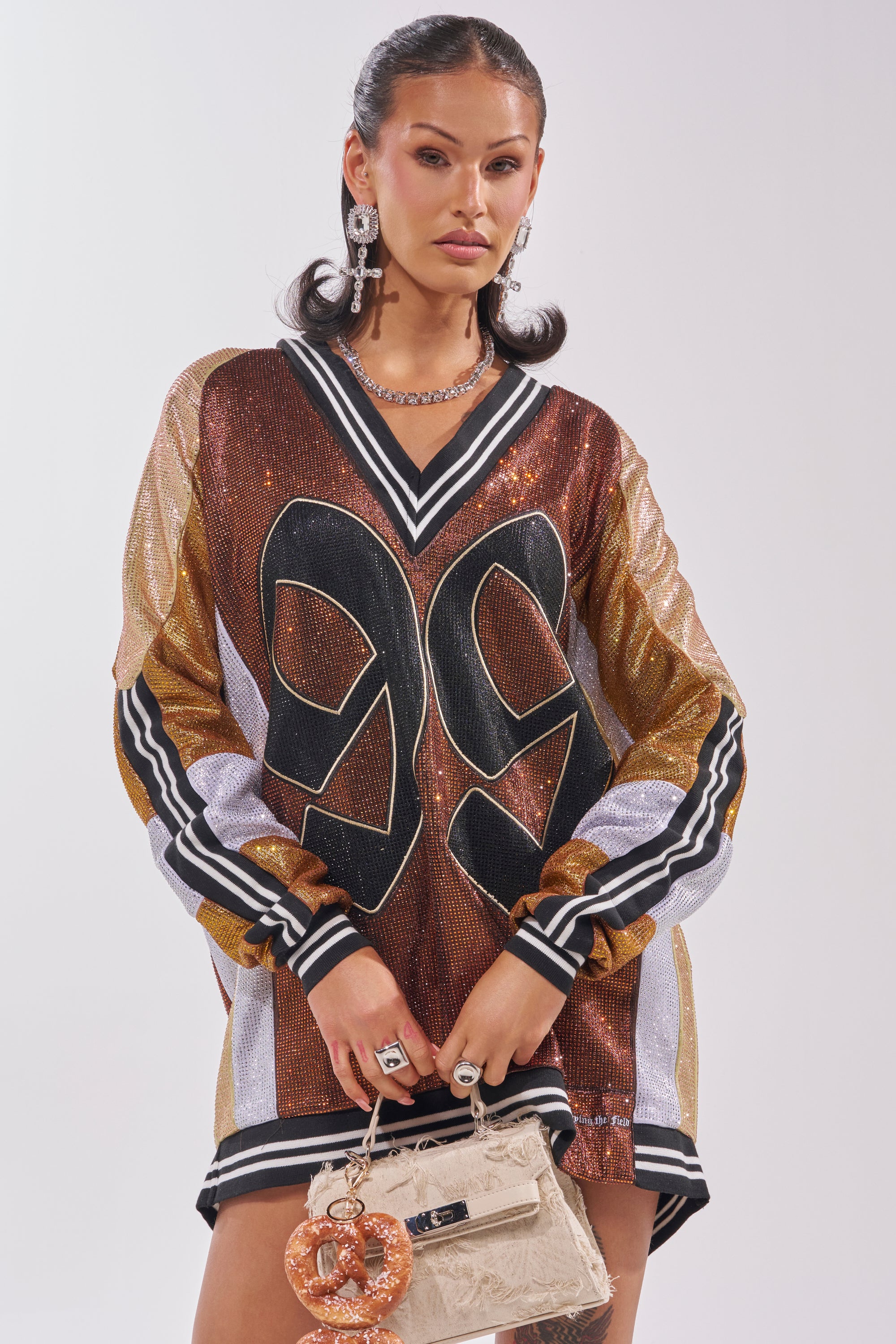 A woman models the RYAN Colorblock Rhinestone Jersey Dress in cocoa, featuring sparkling rhinestones and colorblocked metallic tones. She completes the look with statement jewelry and a beige embellished handbag. 4