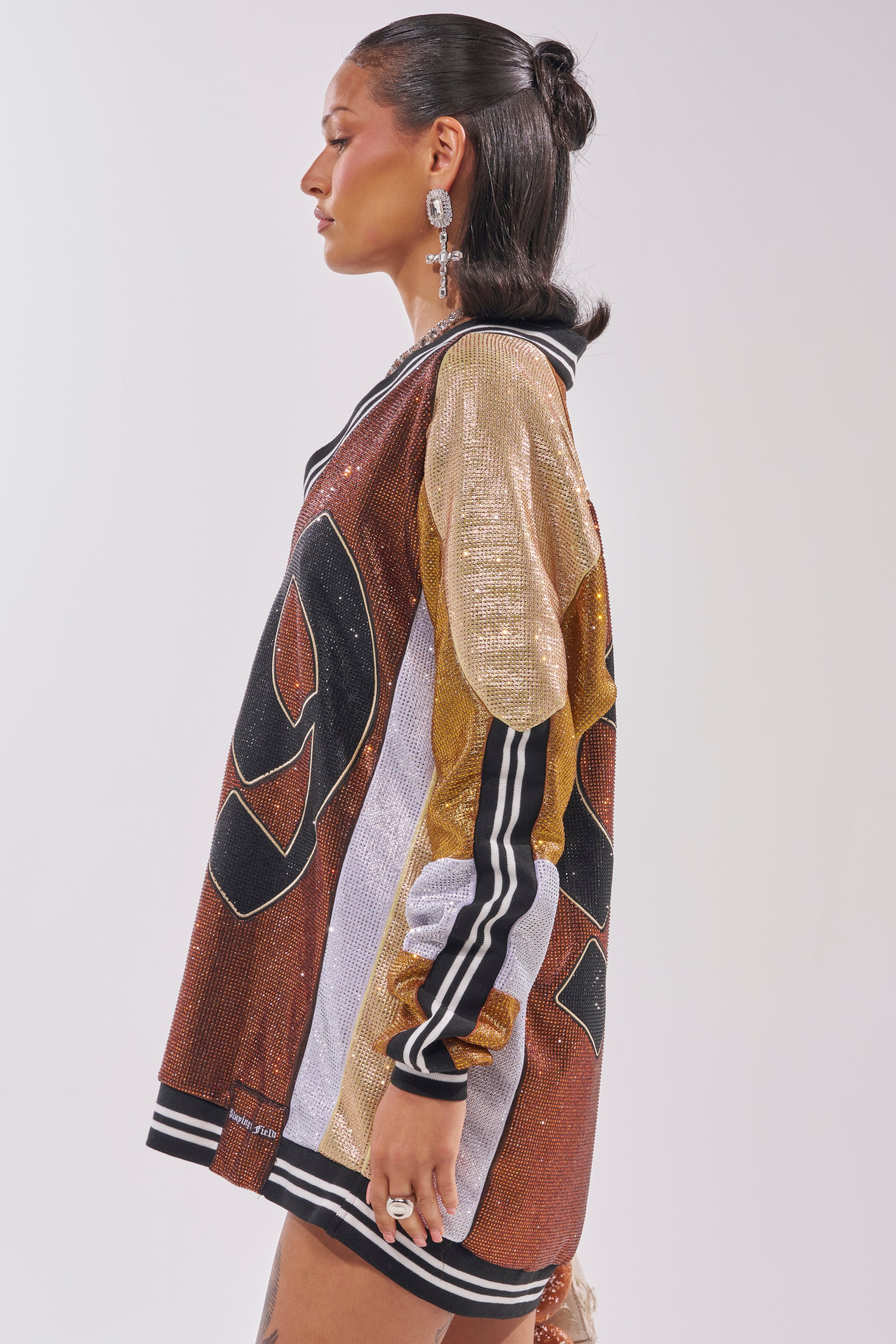 A woman in profile models the RYAN COLORBLOCK RHINESTONE JERSEY DRESS IN COCOA, featuring sparkling gold, bronze, and silver panels with bold black numbers. She has sleek shoulder-length hair and statement earrings. 5