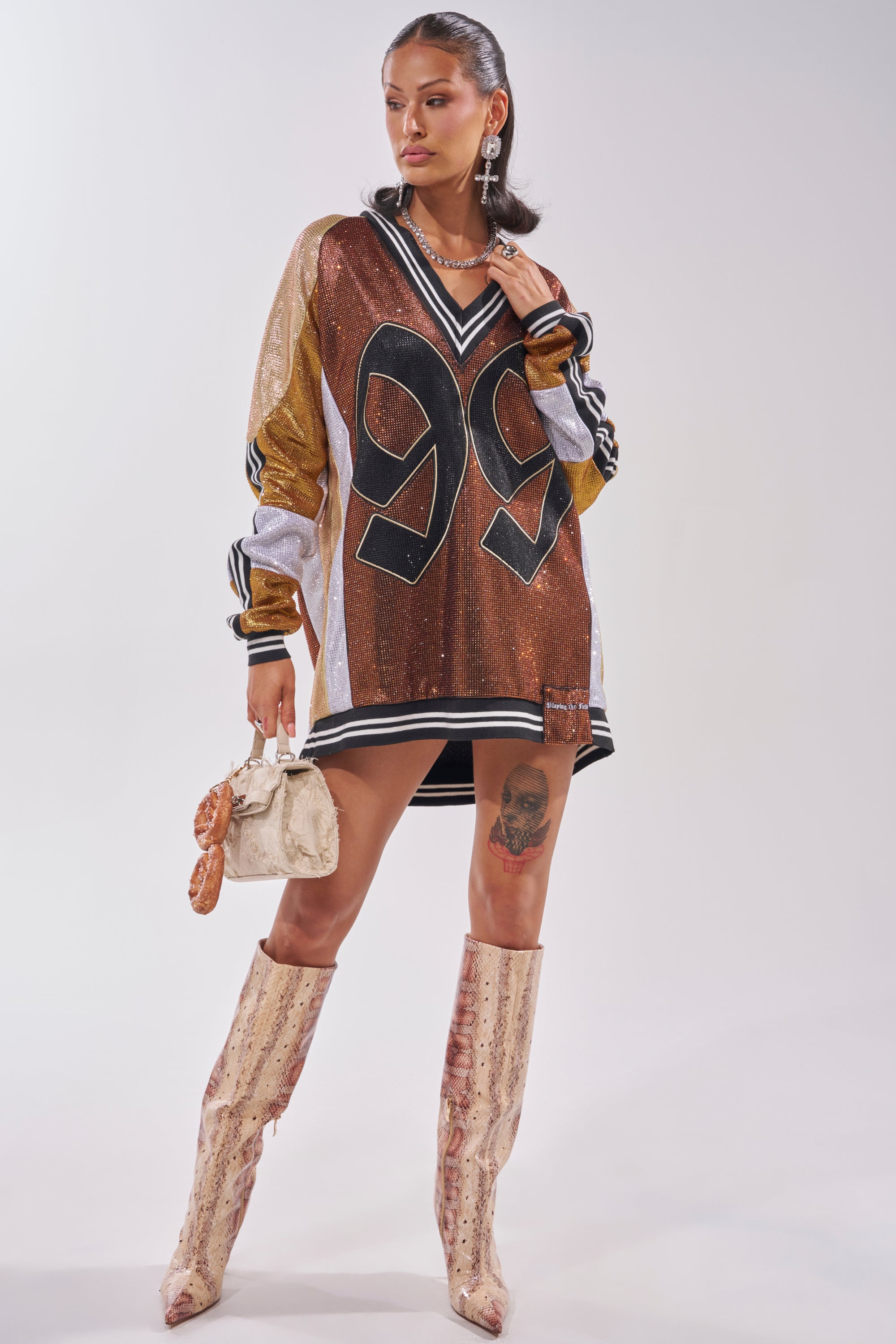 A woman exudes confidence in the RYAN Colorblock Rhinestone Jersey Dress in Cocoa, accessorized with a beige handbag, statement cross earrings, tall patterned boots, and a bold thigh tattoo—an edgy streetwear-inspired look. 7