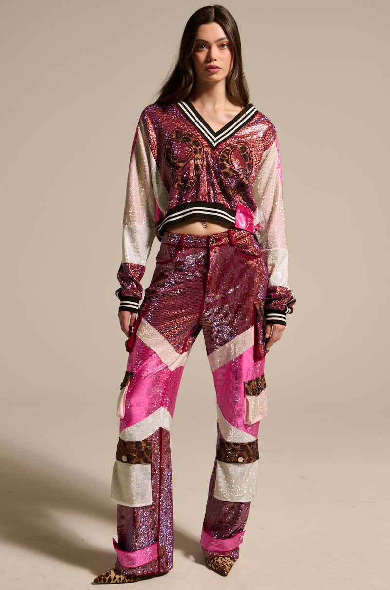 A woman faces forward in ONLY THE COOLEST RHINESTONE PANT, featuring a high-rise fit, chevron and leopard print accents, shiny pink, white and burgundy hues, flared legs, and a glittery finish. Long-sleeved top has striped cuffs.