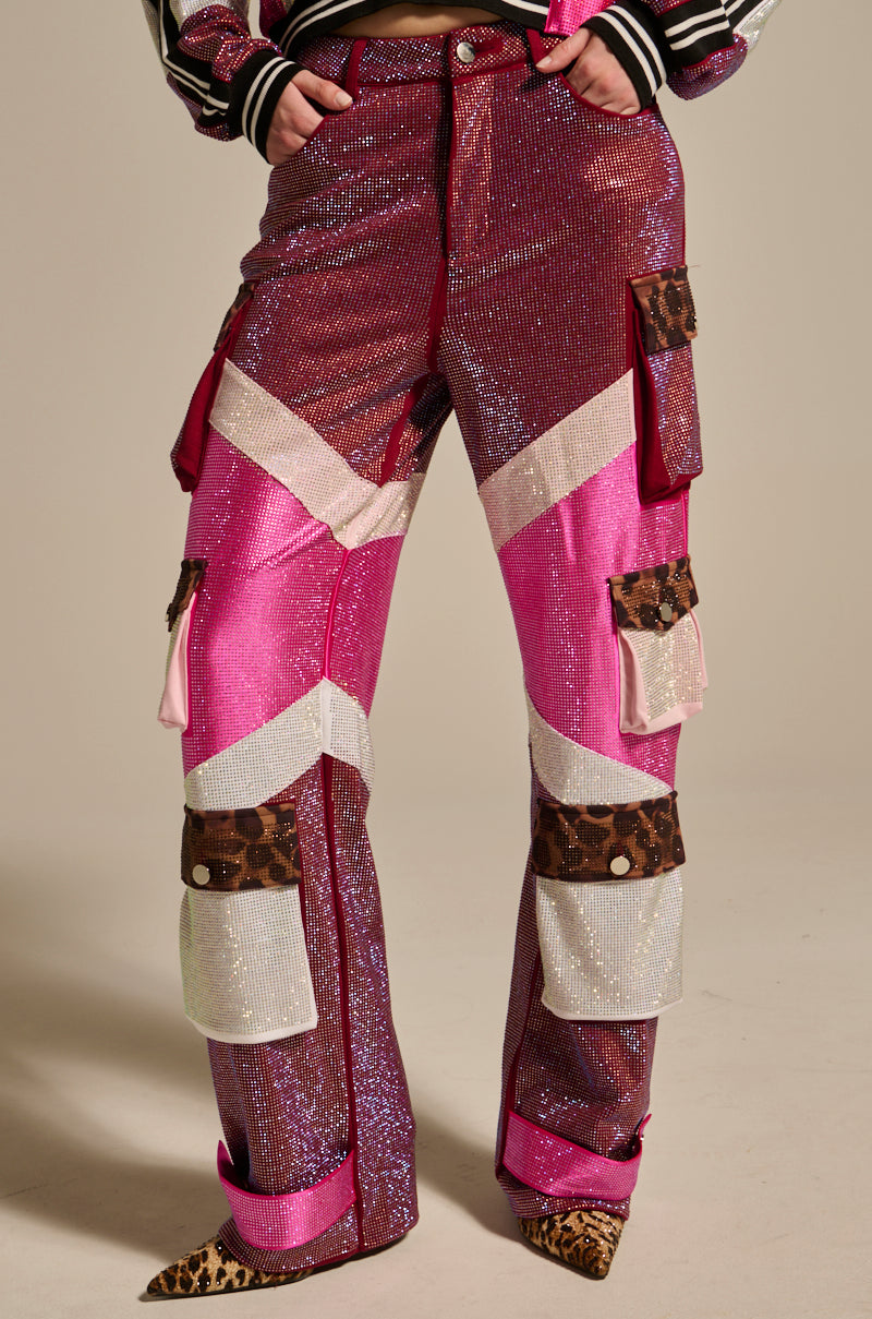 A person wears the ONLY THE COOLEST RHINESTONE PANT—glittery pink and purple high-rise flare cargo pants with white and brown chevron stripes, leopard print patches, and cargo pockets—standing with hands in pockets on a neutral background.