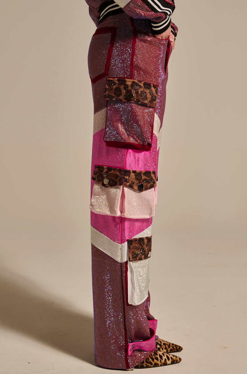 A person stands sideways in ONLY THE COOLEST RHINESTONE PANT—high-rise, shiny pink with silver stripes, animal print patches, white and pink accents—paired with a patterned top and leopard-print shoes against a neutral backdrop.
