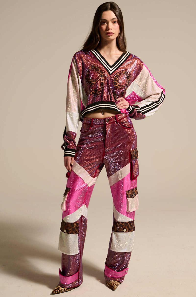 A woman stands confidently, wearing a glittery, colorful outfit with the ONLY THE COOLEST RHINESTONE PANT featuring shiny patterns and patchwork details, paired with a matching pink, purple, and white long-sleeve top and animal print shoes.