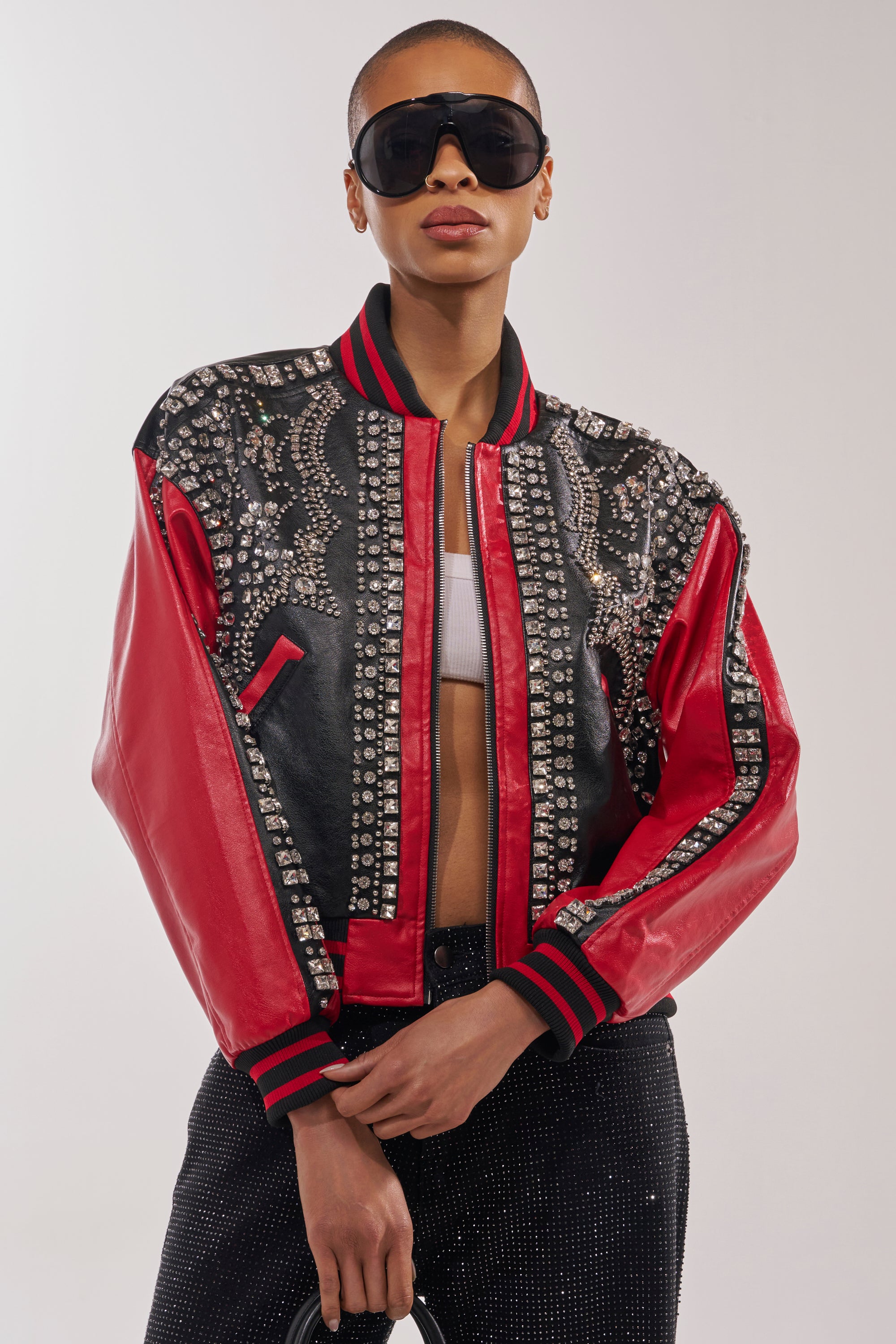 A person stands confidently against a plain background wearing the STOLE THE SHOW EMBELLISHED BOMBER, large black sunglasses, a white crop top, and black pants. 0
