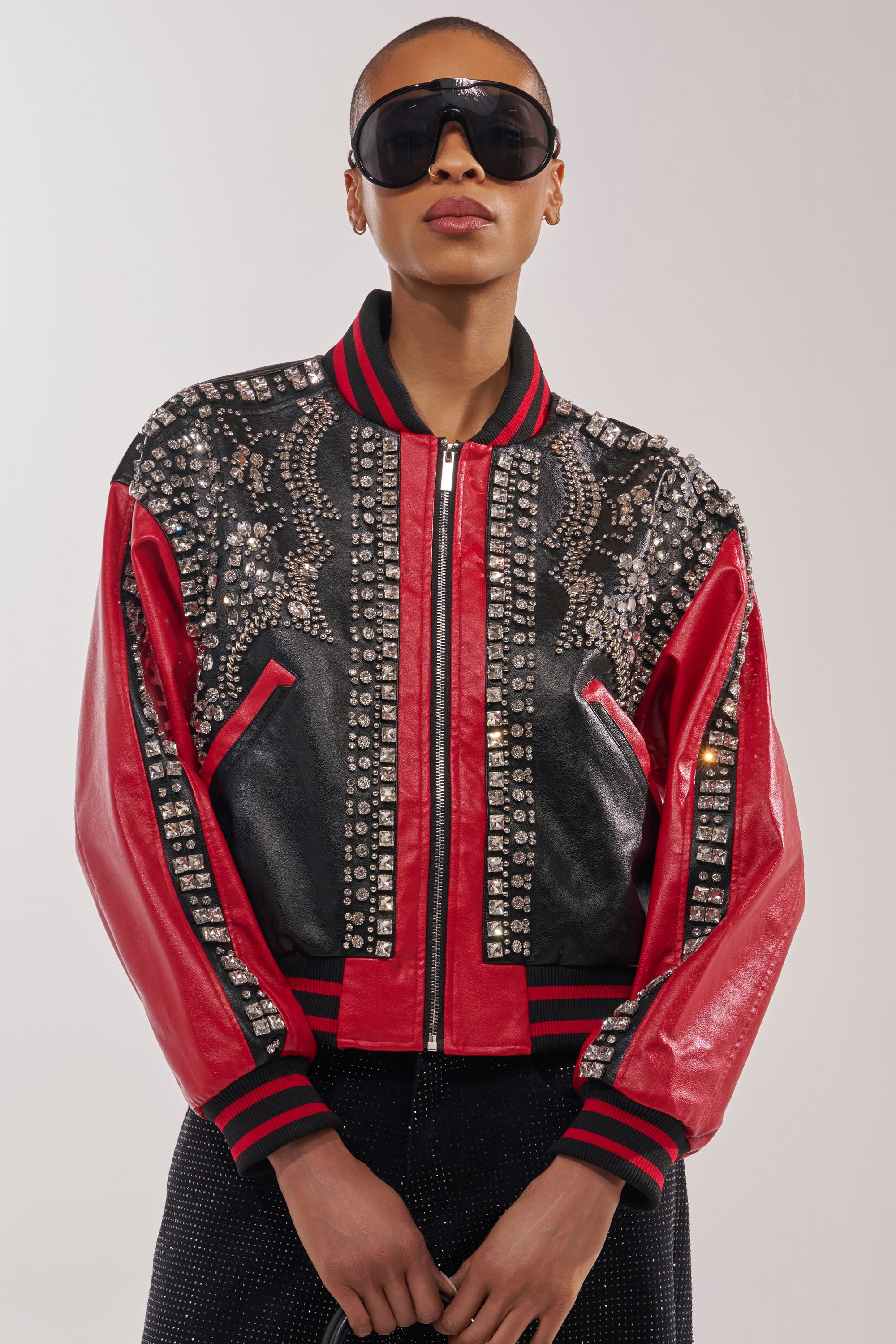 A person wearing large dark sunglasses and the STOLE THE SHOW EMBELLISHED BOMBER, featuring bold rhinestones, silver studs, and intricate details, stands against a plain light background. 3