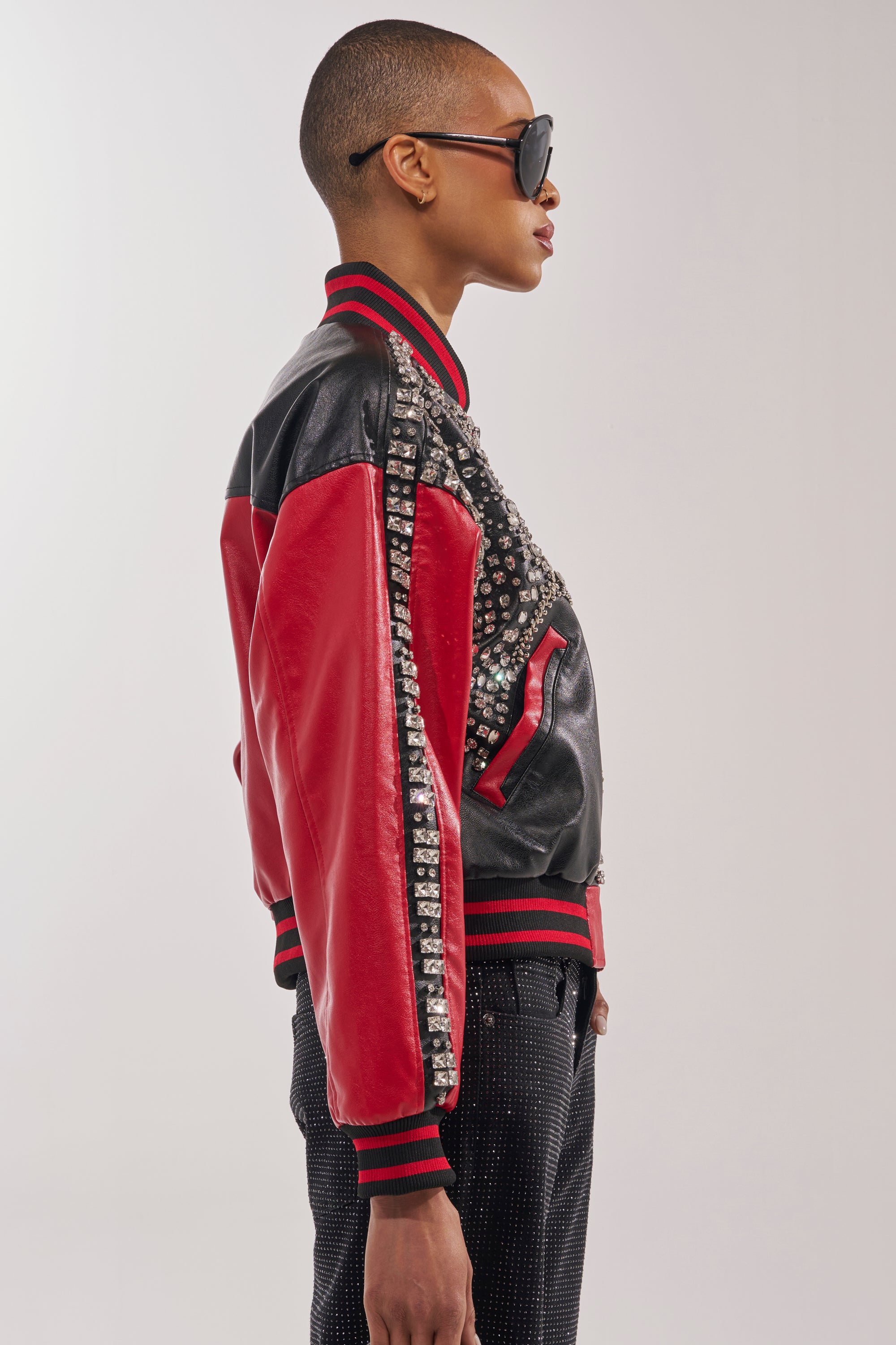 A person in profile stands against a plain light background wearing the STOLE THE SHOW EMBELLISHED BOMBER in black and red with studs, large sunglasses, and dark pants. 4