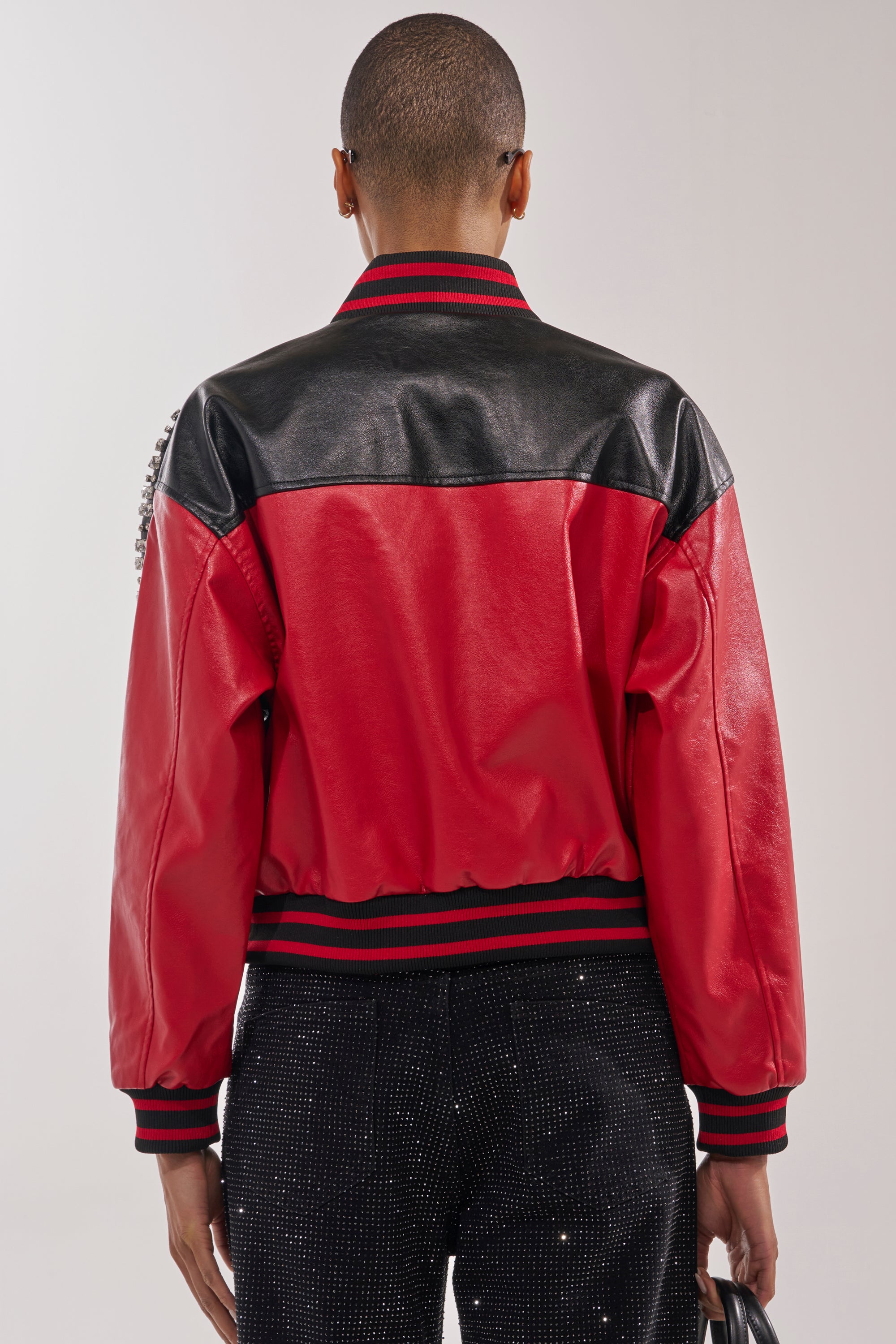 A person with short hair is seen from behind wearing the STOLE THE SHOW EMBELLISHED BOMBER—a red and black faux leather jacket with striped cuffs and collar—paired with black pants featuring small white dots. 5