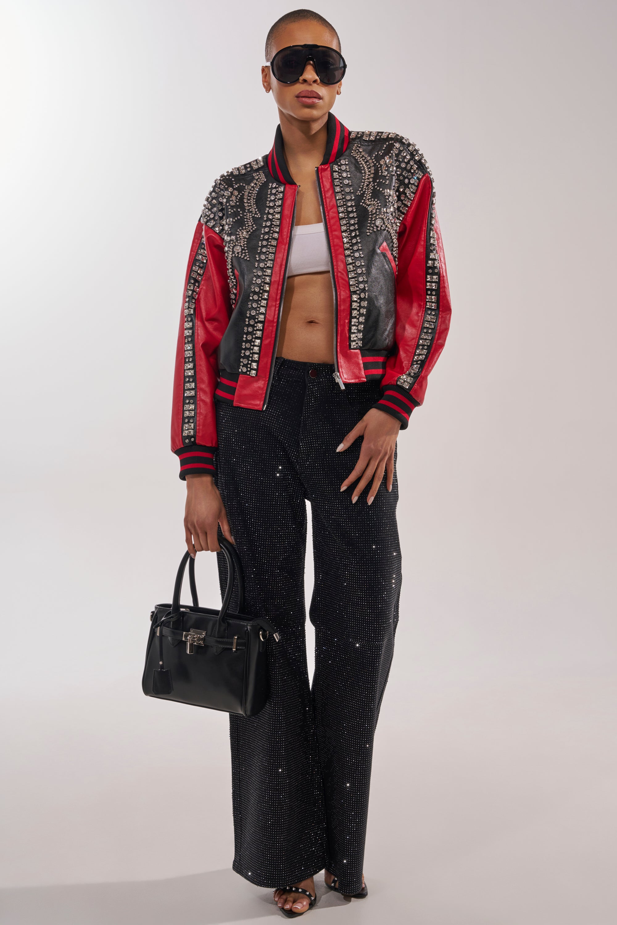 A stylish person holds a black handbag while wearing sunglasses, the STOLE THE SHOW EMBELLISHED BOMBER in red and black, a white crop top, sparkly black pants, and open-toe heels against a plain background. 6