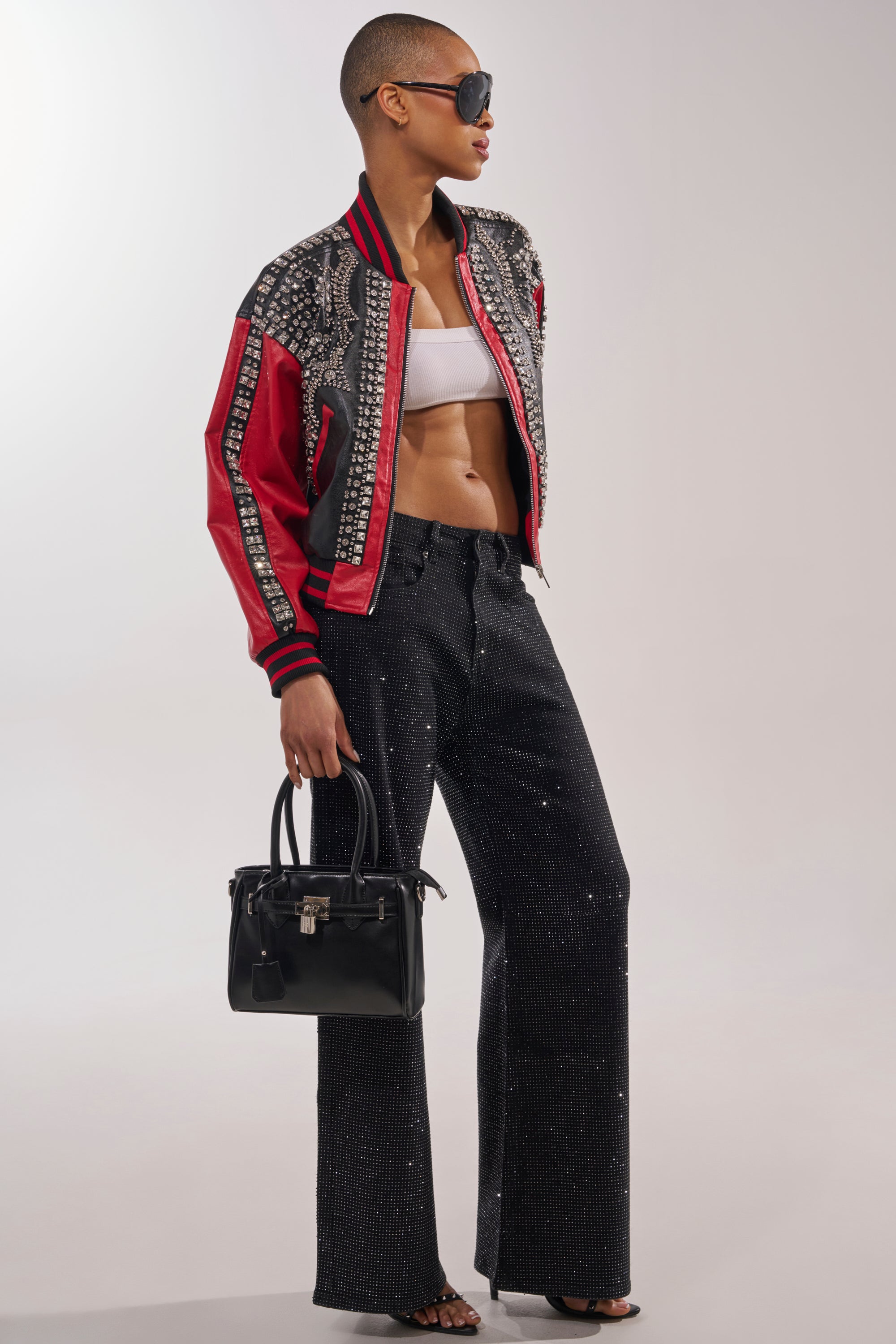 Wearing sunglasses, the person sports the STOLE THE SHOW EMBELLISHED BOMBER in black and red over a white bandeau top, paired with black sparkling wide-leg pants, black sandals, and a black handbag against a plain backdrop. 7