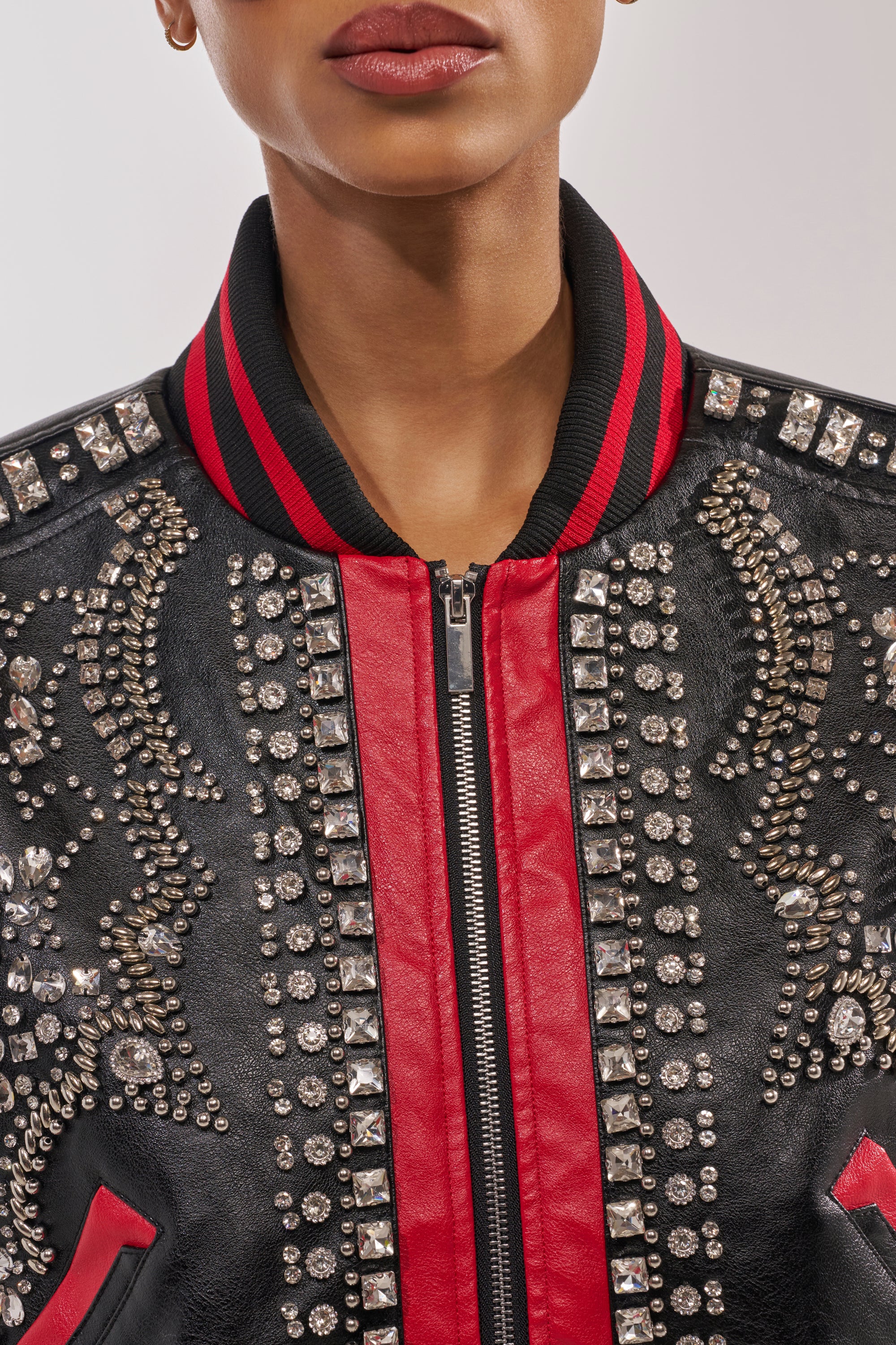 A person wears the STOLE THE SHOW EMBELLISHED BOMBER, a black and red faux leather jacket with silver studs, rhinestones, and a black collar with red stripes. Their face is partially visible from the nose down. 8