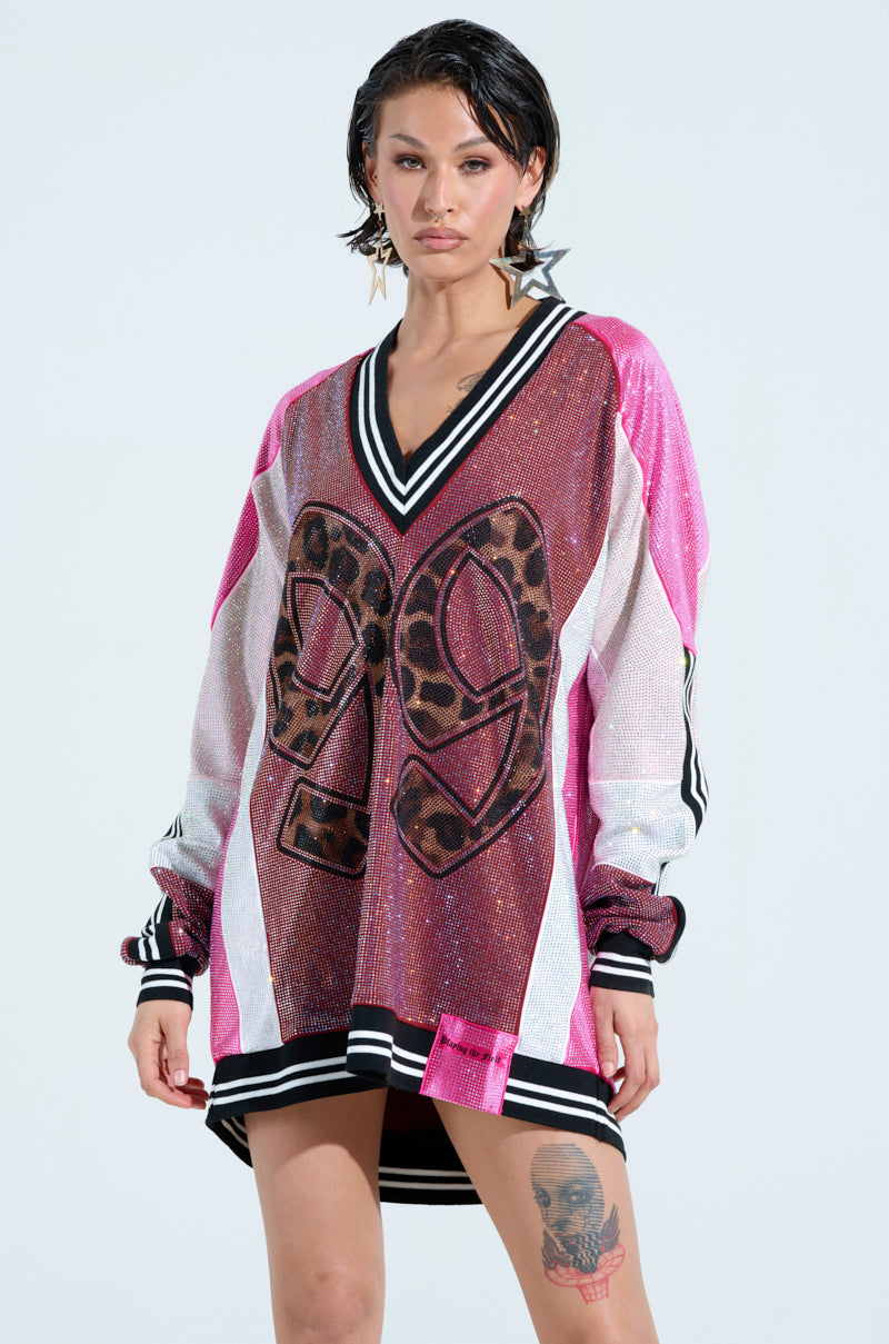 A model wears the ONLY THE COOLEST RHINESTONE JERSEY DRESS, featuring a long, oversized leopard print with "29" on the front. Black and white striped sleeves enhance her sporty look. Her shoulder-length hair adds elegance against a plain background.