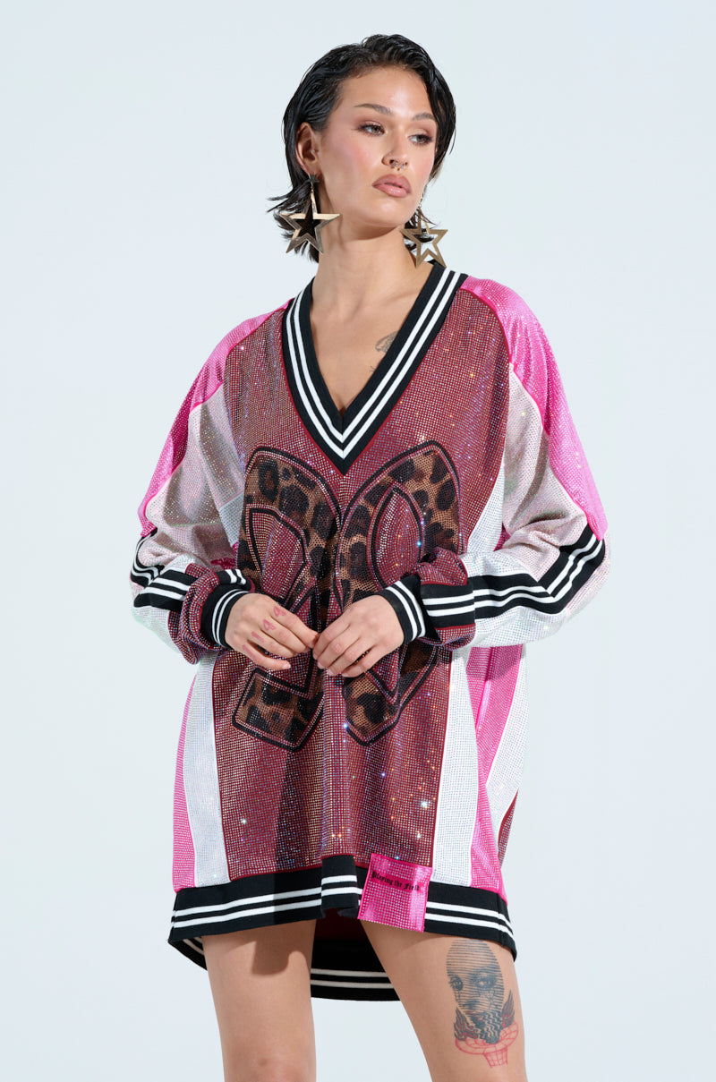 A person in an ONLY THE COOLEST RHINESTONE JERSEY DRESS, featuring pink sequins with black and white stripes and a letter on the front. They sport short hair, star-shaped earrings, a visible thigh tattoo, creating an 'oversized sporty dress' vibe against a plain light-colored backdrop.