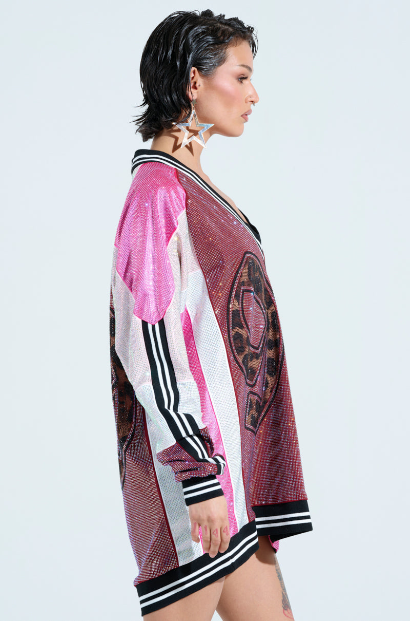 A person in an oversized, colorful jersey dress with large patterns and star-shaped earrings reminiscent of sportswear. The dress features pink, white, and black stripes. They have short, dark hair styled wet-look and stand against a plain background.