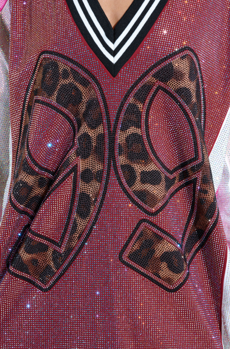 Someone is dressed in the ONLY THE COOLEST RHINESTONE JERSEY DRESS, a sporty oversized piece in maroon glitter fabric with leopard print "99" numerals outlined in black and a bold black and white striped neckline.
