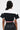 A person with long dark hair wears the OFFICIAL DUTY CROPPED BUTTON DOWN TOP, featuring a color block pattern with red and white trim, paired with black high-waisted pants. They face away from the camera against a neutral background.