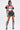 A person confidently wears the OFFICIAL DUTY CROPPED BUTTON DOWN TOP, styled with a black/red color block pattern, black faux leather shorts with a chain detail, and knee-high black boots featuring white lace-up accents, all set against a plain white background.