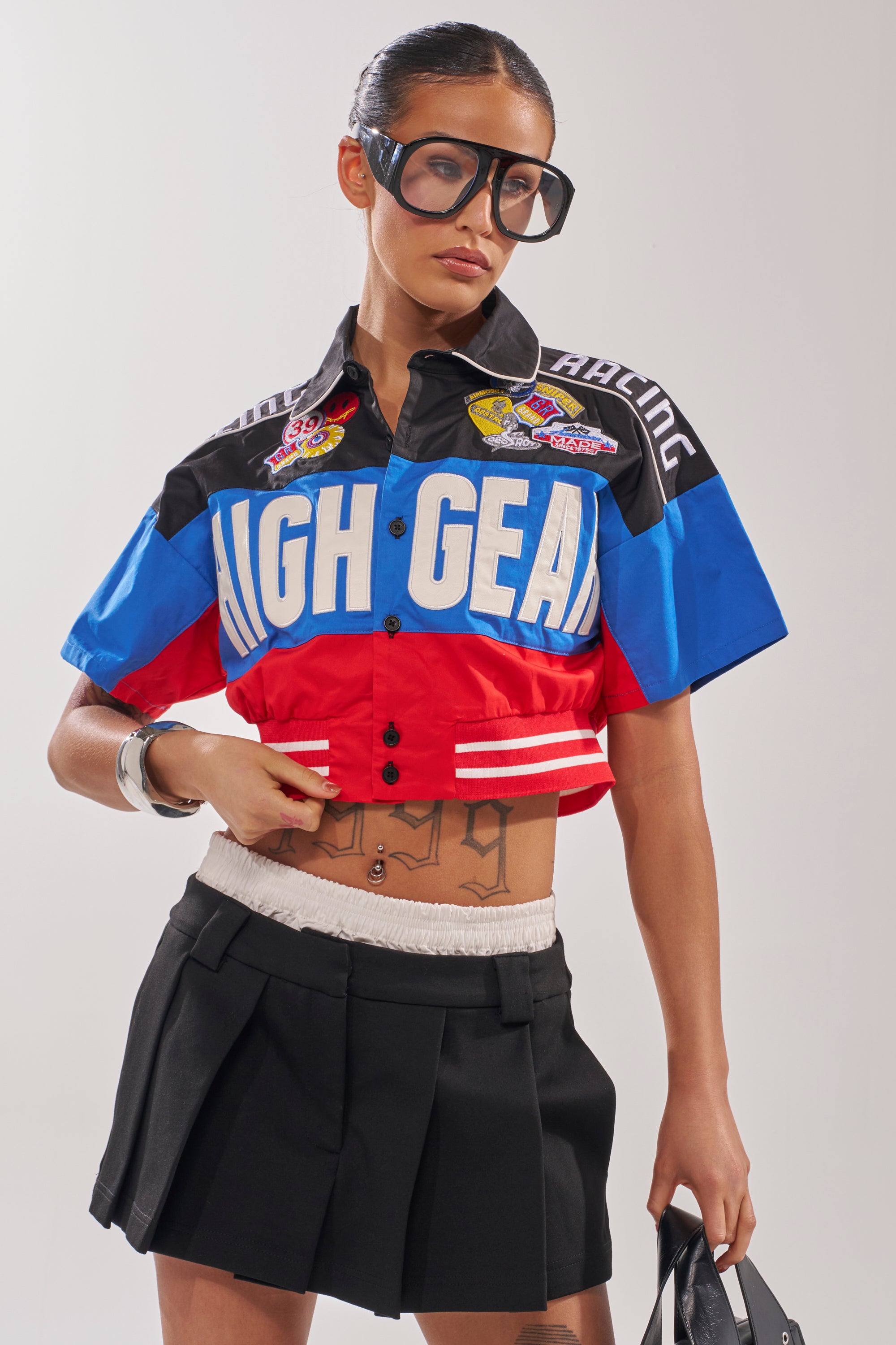 A woman in oversized sunglasses and the HIGH GEAR CROPPED BUTTON DOWN, paired with a black pleated mini skirt, stands confidently with one hand on her hip, showing off her midriff and tattoos in bold streetwear style. 1