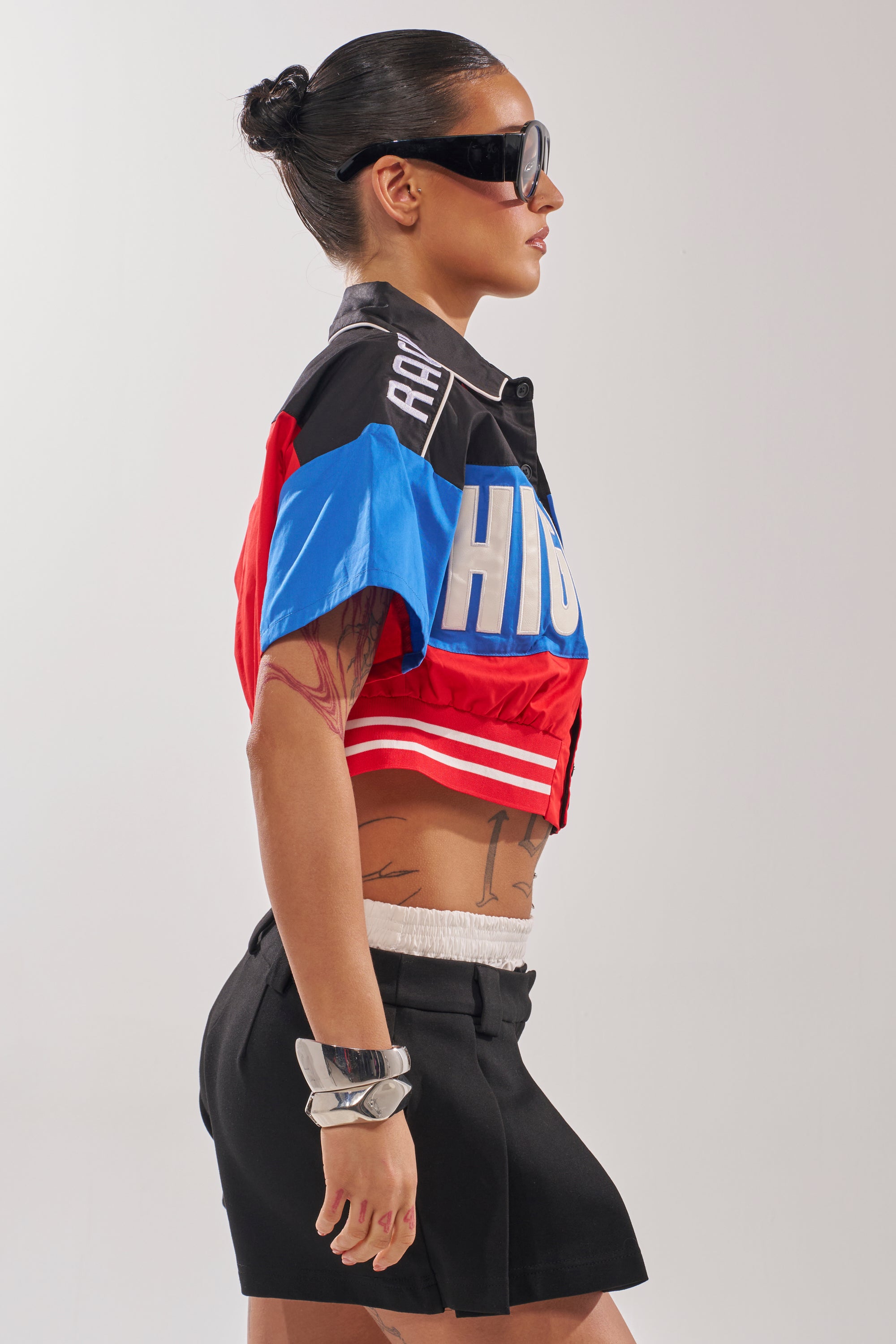 A woman with dark hair in a bun, large sunglasses, and visible tattoos wears the HIGH GEAR CROPPED BUTTON DOWN streetwear jacket with black shorts and a silver bracelet, standing in profile against a plain background. 2