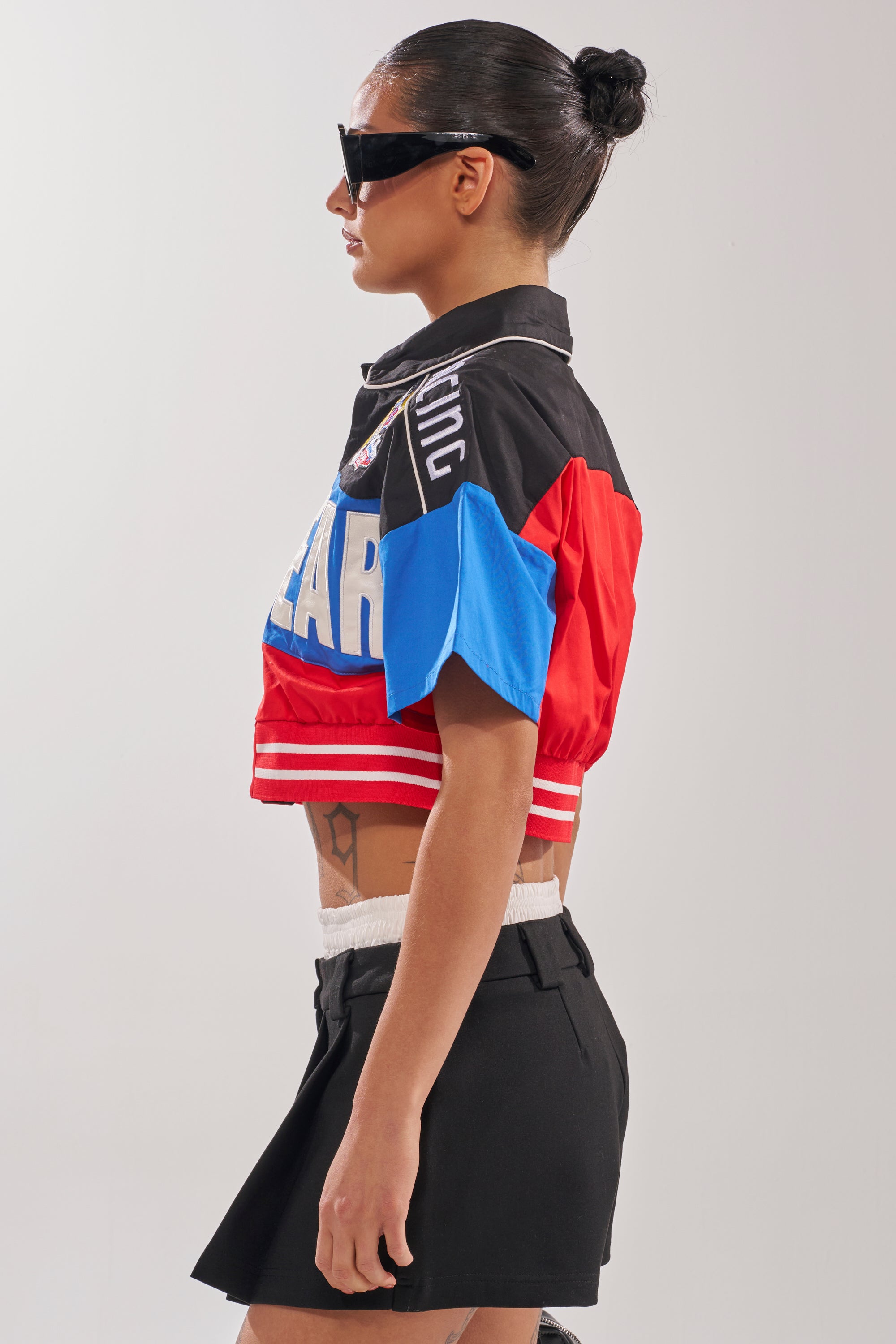 A woman with dark hair in a bun wears black sunglasses, a HIGH GEAR CROPPED BUTTON DOWN, and a black mini skirt, standing in profile against a plain light background. 3